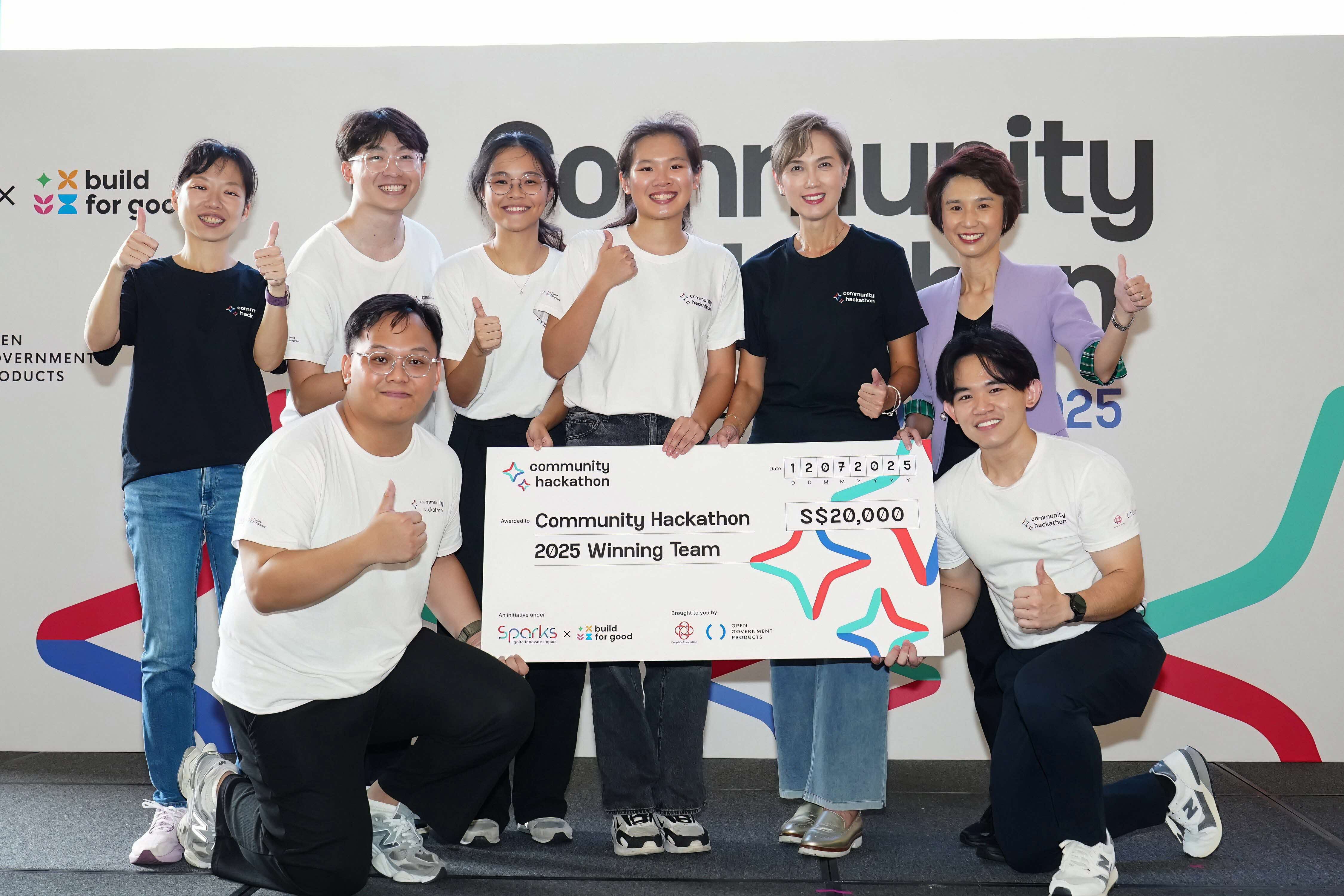 Eight people celebrating at a Community Hackathon event, with six standing in the back row giving thumbs up and two kneeling in front holding a large ceremonial cheque for S$20,000 made out to the "Community Hackathon 2025 Winning Team." Most participants wear white t-shirts with event branding. The backdrop shows "Community Hackathon" branding with "build for good" and "Open Government Products" logos visible on the left side.