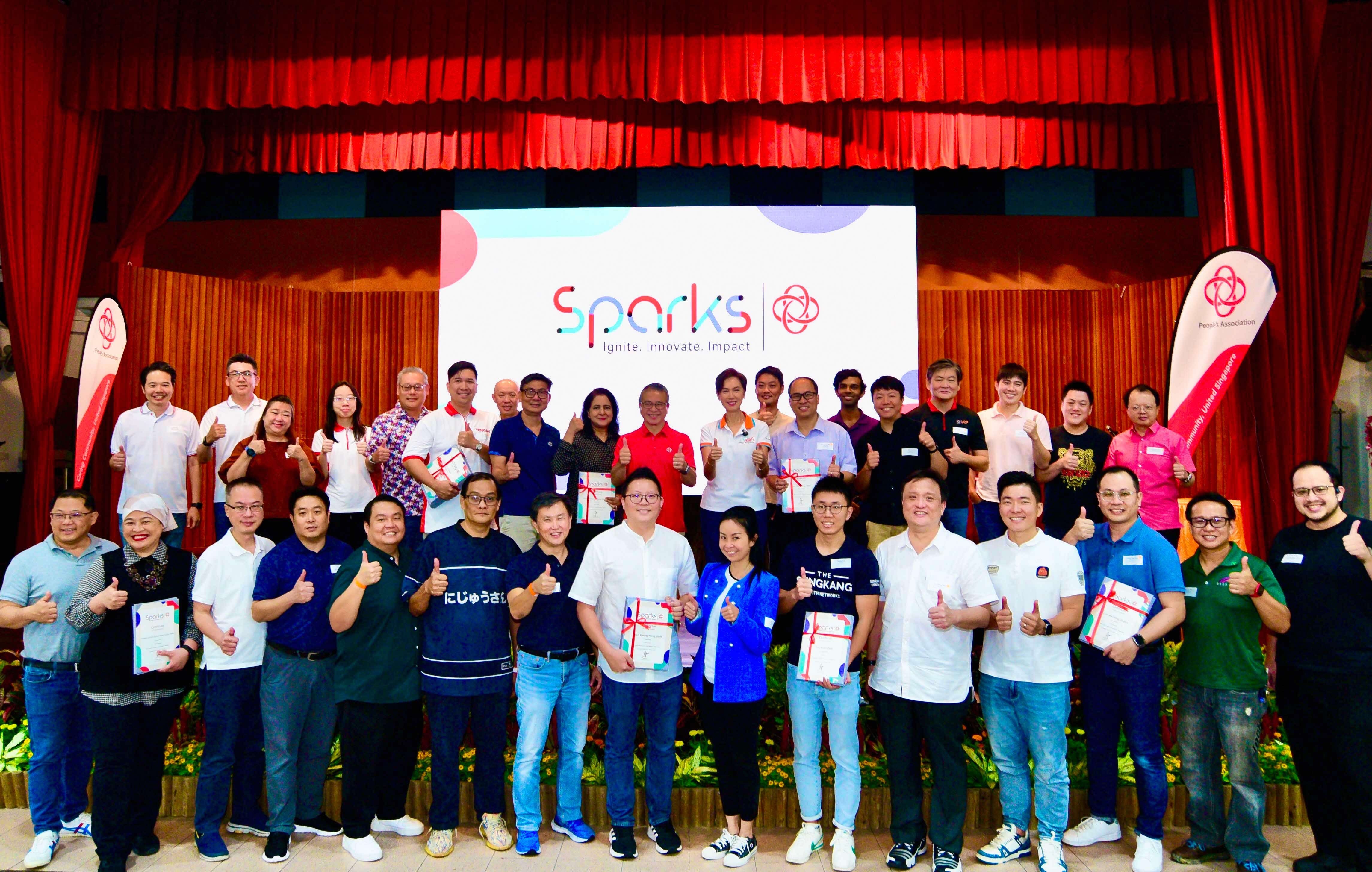 A large group photo of approximately 35 people at a Sparks event, arranged in two rows on a stage with red curtains. Participants are giving thumbs up gestures and many are holding certificates or awards. Behind them is a large screen displaying the "Sparks - Ignite. Innovate. Impact" logo, with branded banners on either side of the stage.