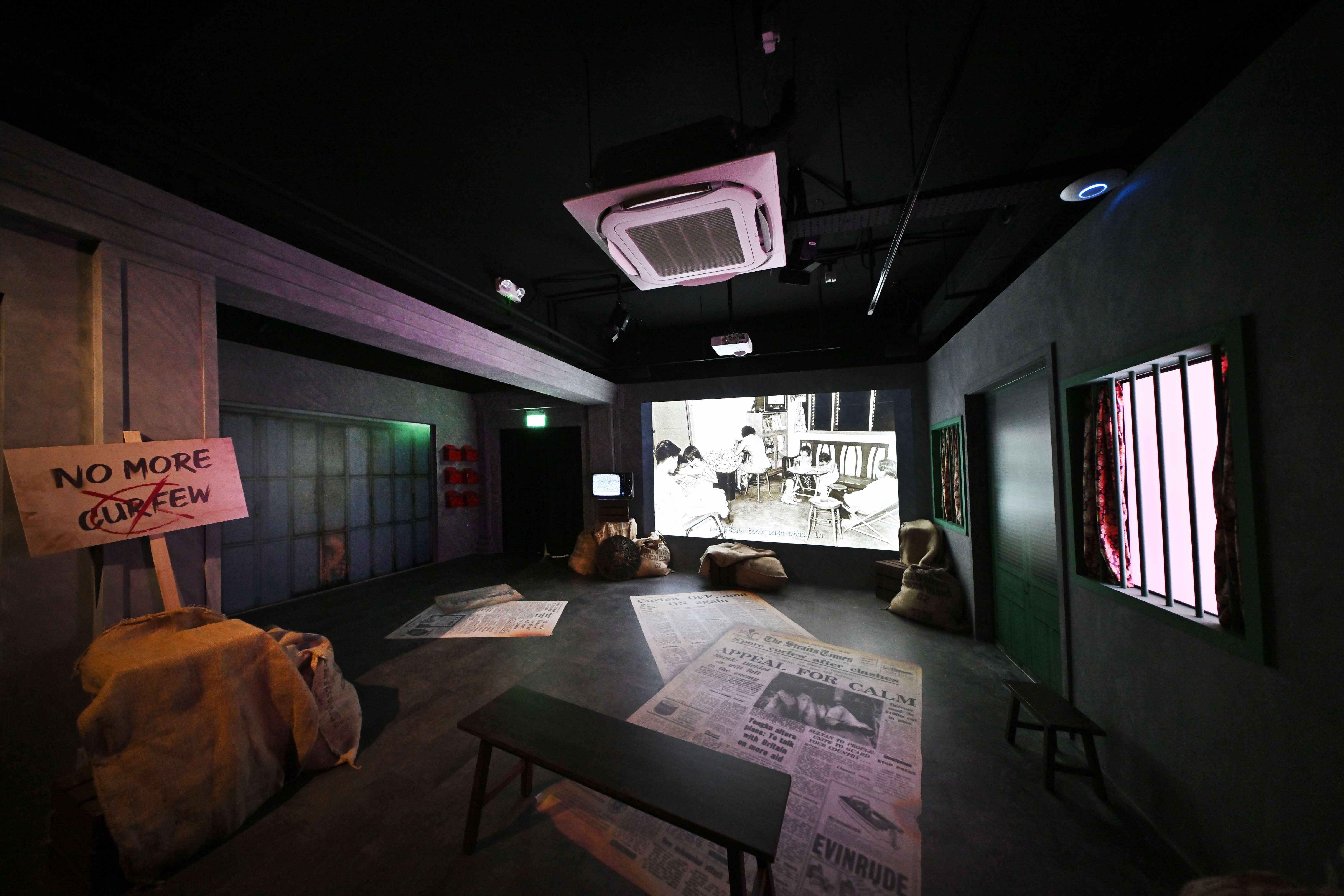 A room with a "No More Curfew" sign, sack bags, a bench, large newspaper pages on the floor, and a vintage photo projection.