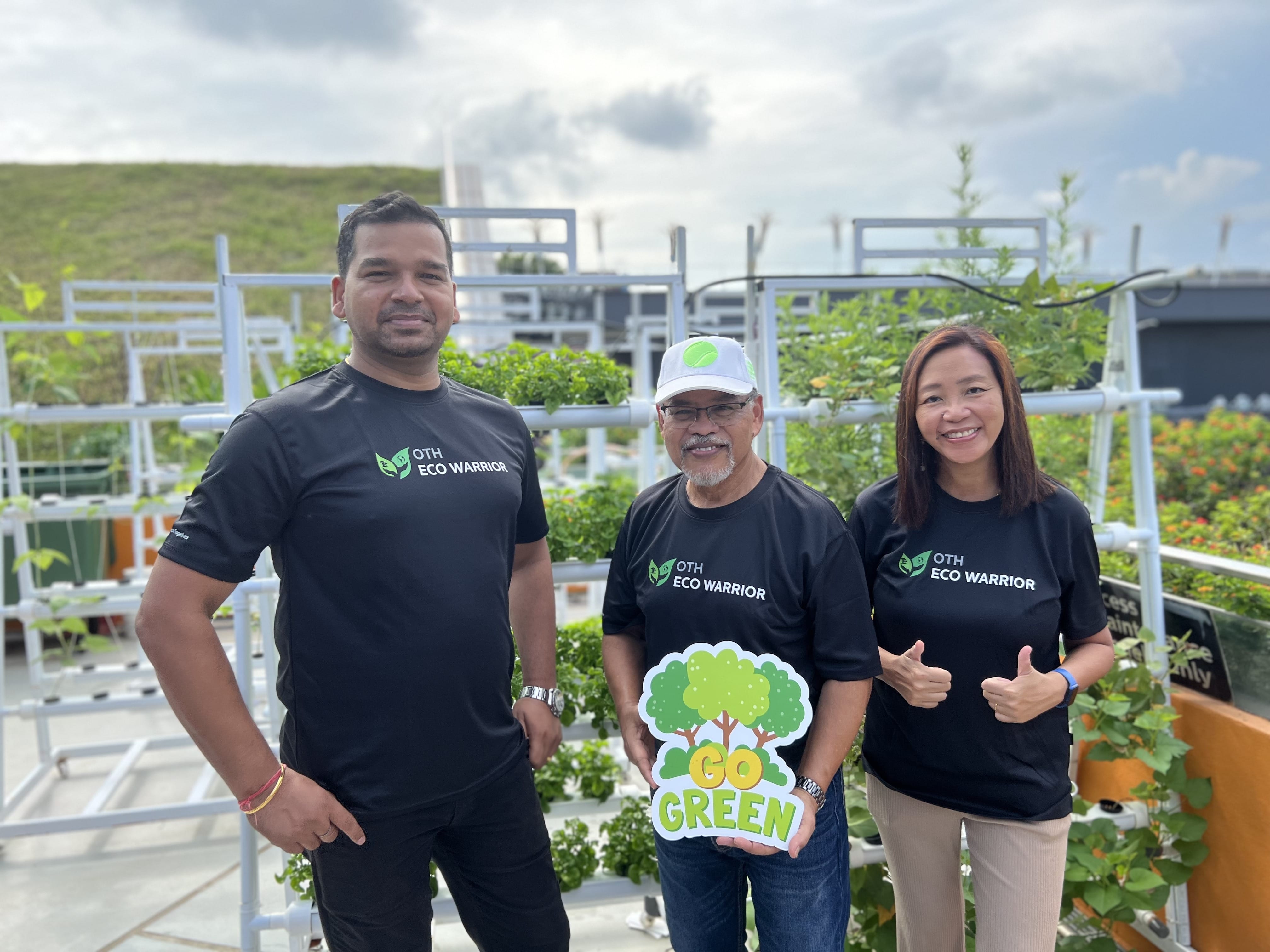 Three volunteers wearing black "OTH Eco Warrior" t-shirts posing together in a rooftop garden, holding a "Go Green" sign with tree logo, surrounded by plants and urban farming setup.