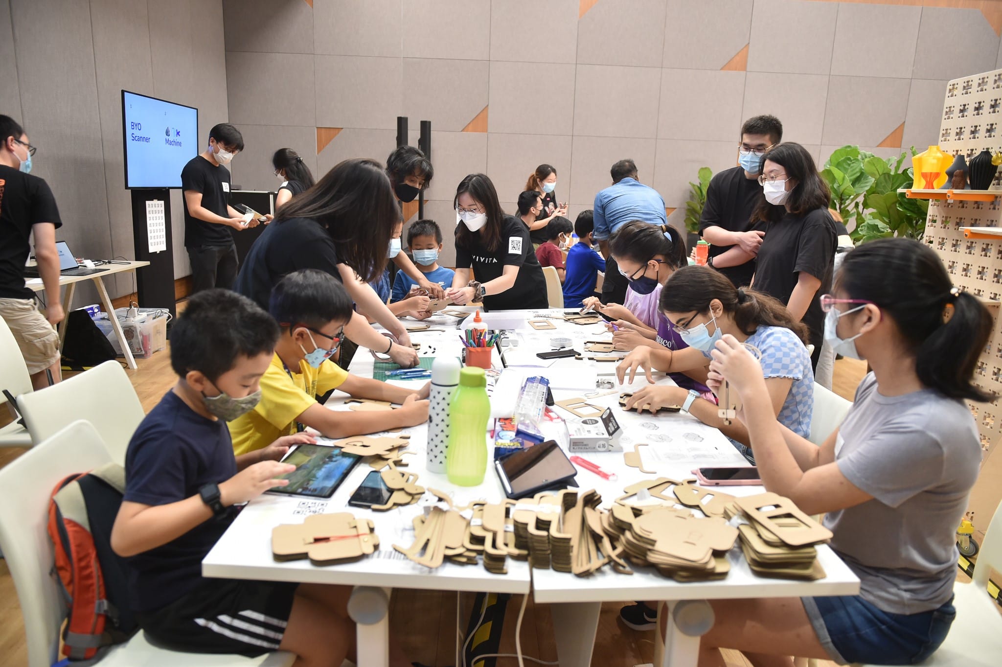 Children and adults are seated around a table, working on a craft project with cardboard materials and tablets.
