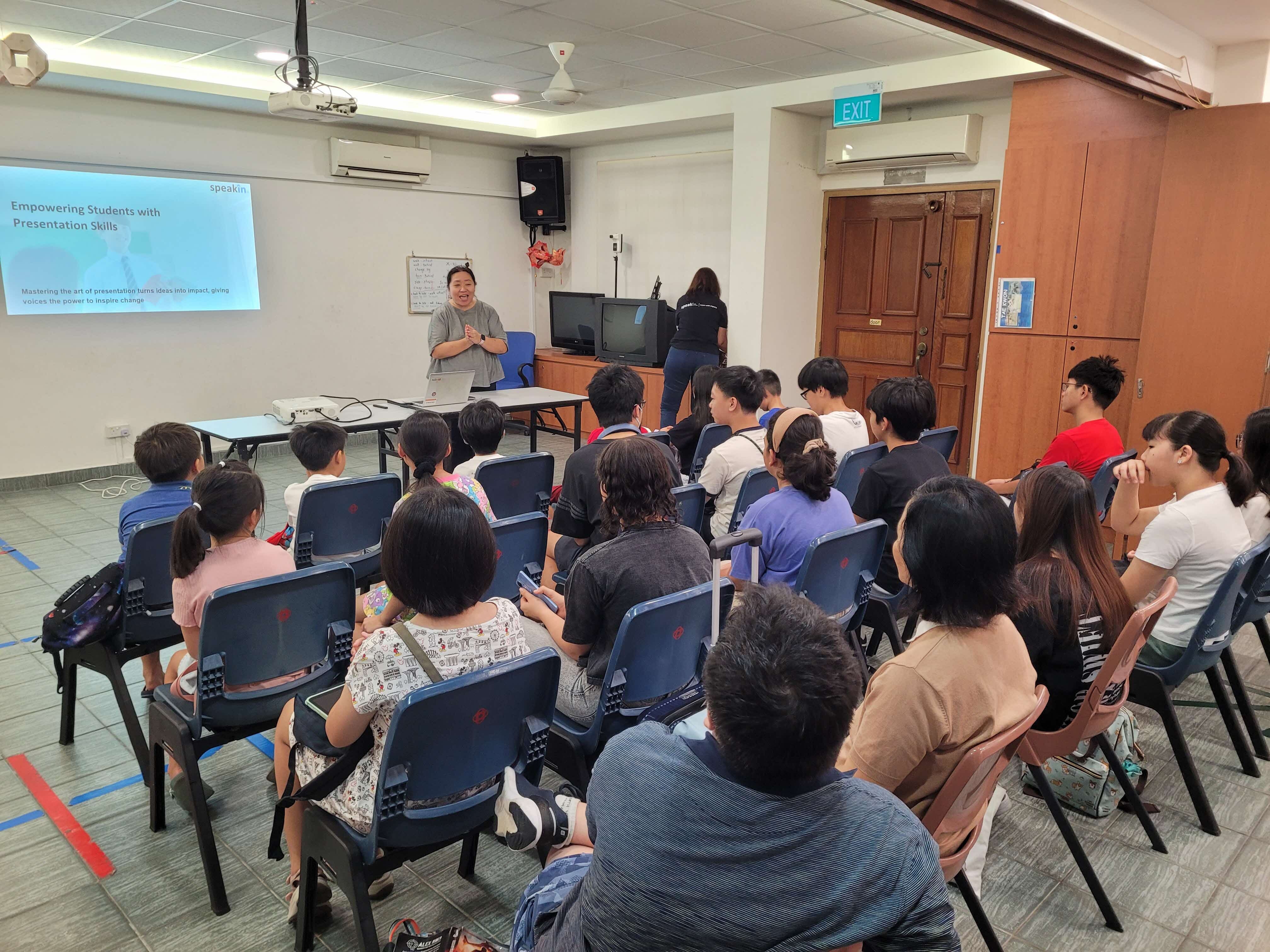  A presentation session with approximately 30 students seated in blue chairs facing a presenter who stands beside a screen displaying "Empowering Students with Presentation Skills" in the same conference room.