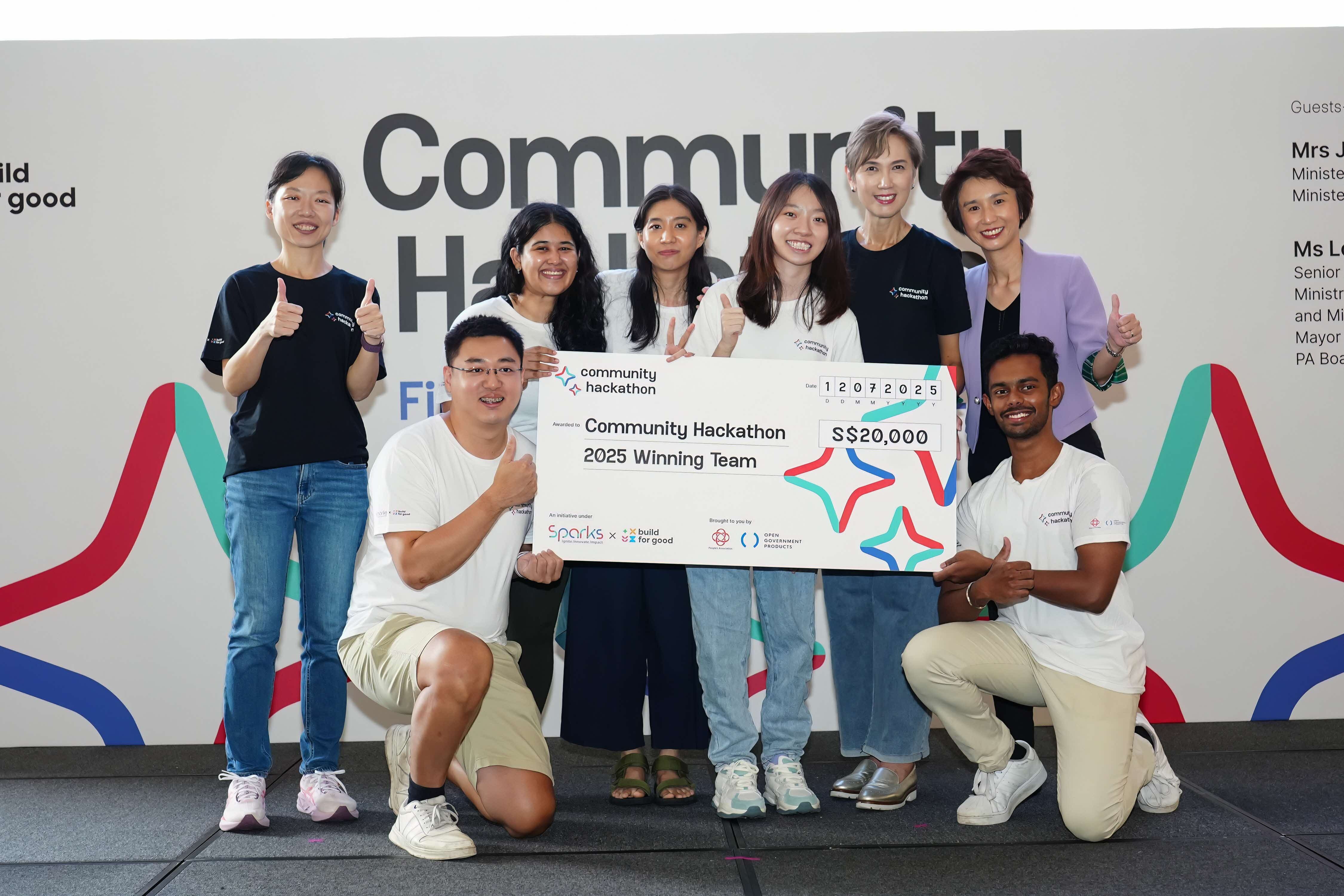 Eight people celebrating at a Community Hackathon event, holding a large ceremonial cheque for S$20,000 made out to the "Community Hackathon 2025 Winning Team." Six people stand in the back row giving thumbs up, while two men kneel in front holding the cheque. They're positioned in front of a wall with "Community Hackathon" branding and colorful curved design elements.
