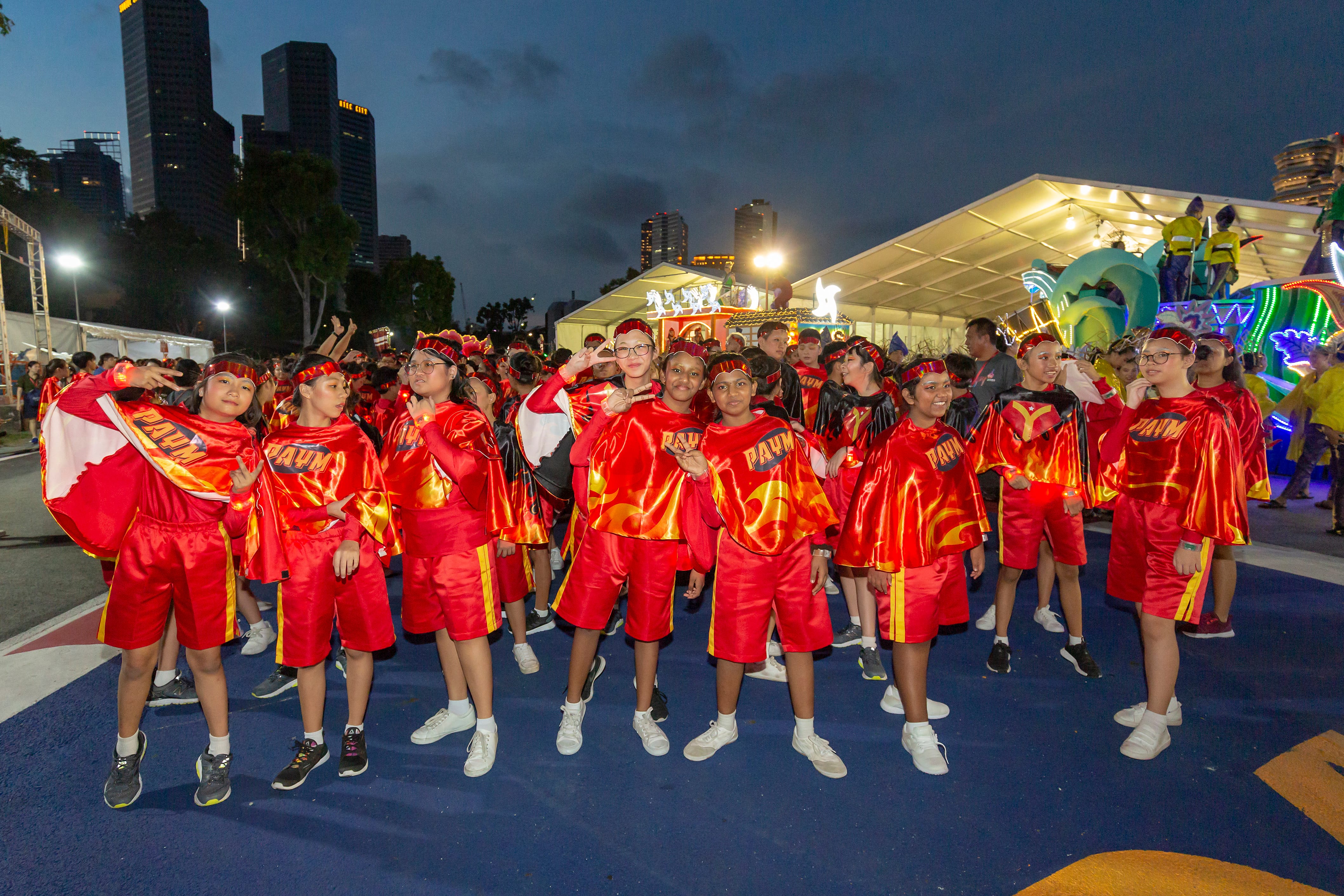 A group wearing red and gold outfits stands together in an outdoor area with illuminated floats and city buildings in the background.