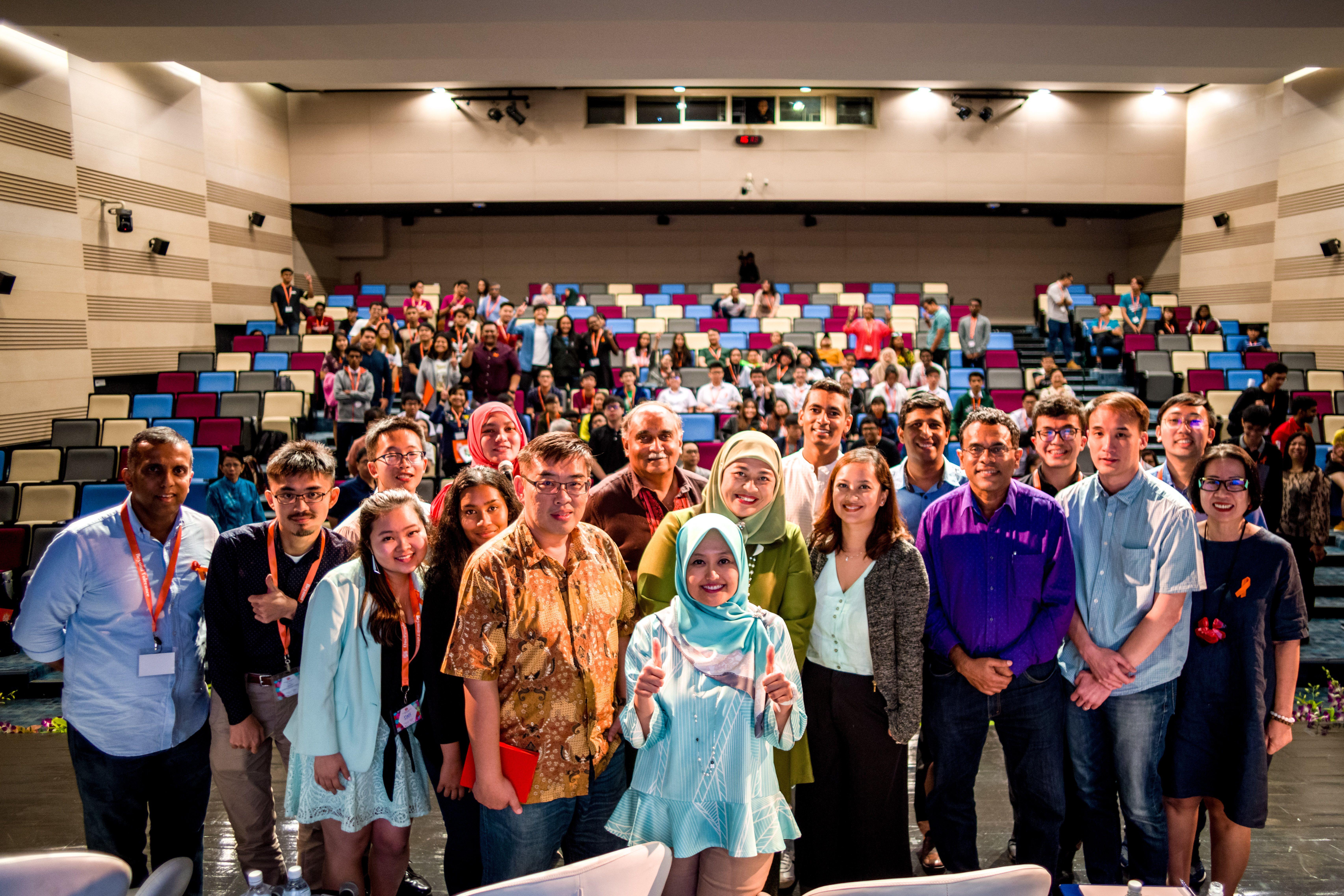 A group of people stand at the front of a packed auditorium with colorful seats arranged in tiers.
