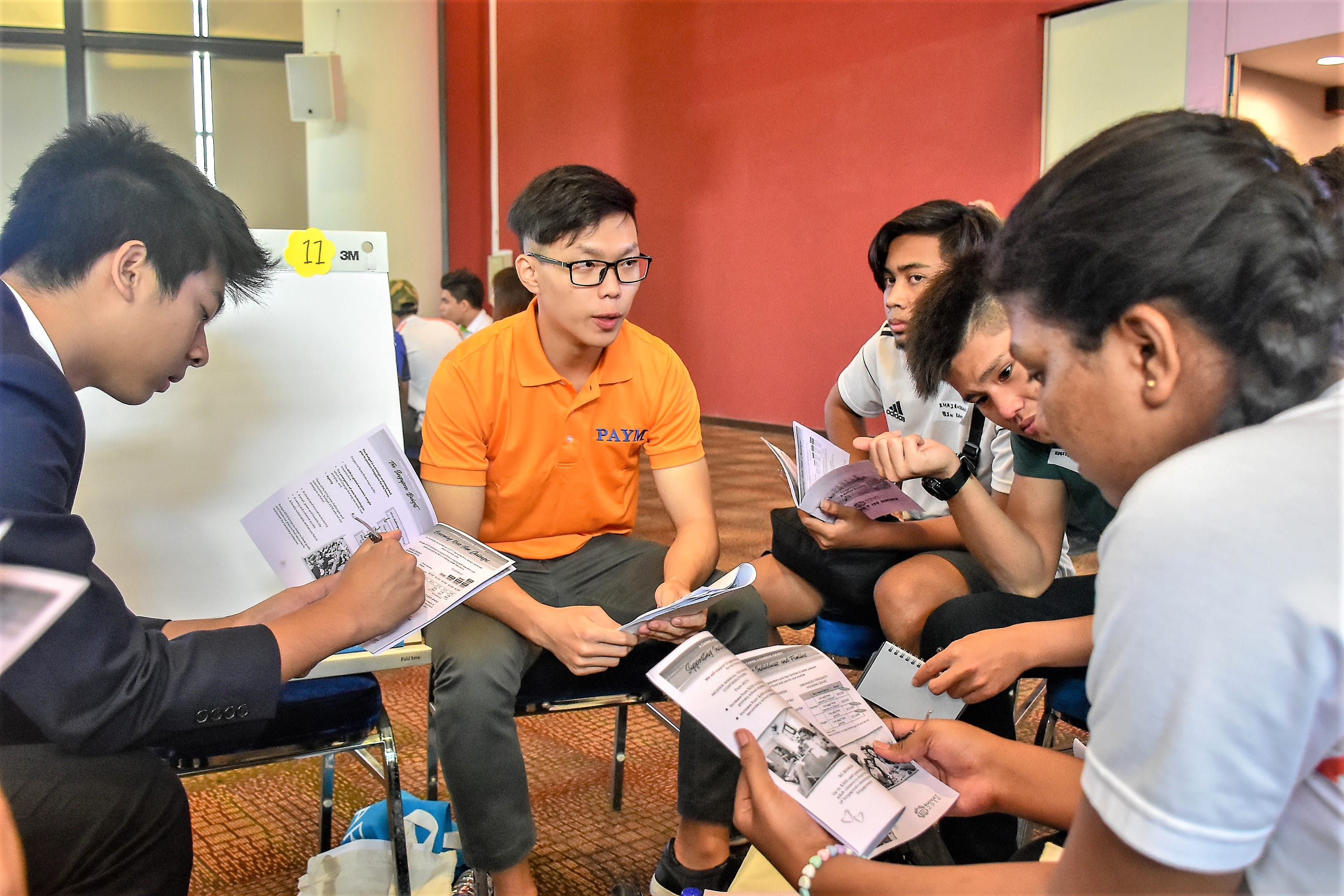 A group of people sitting in chairs, holding and discussing printed materials in a room with red and white walls.