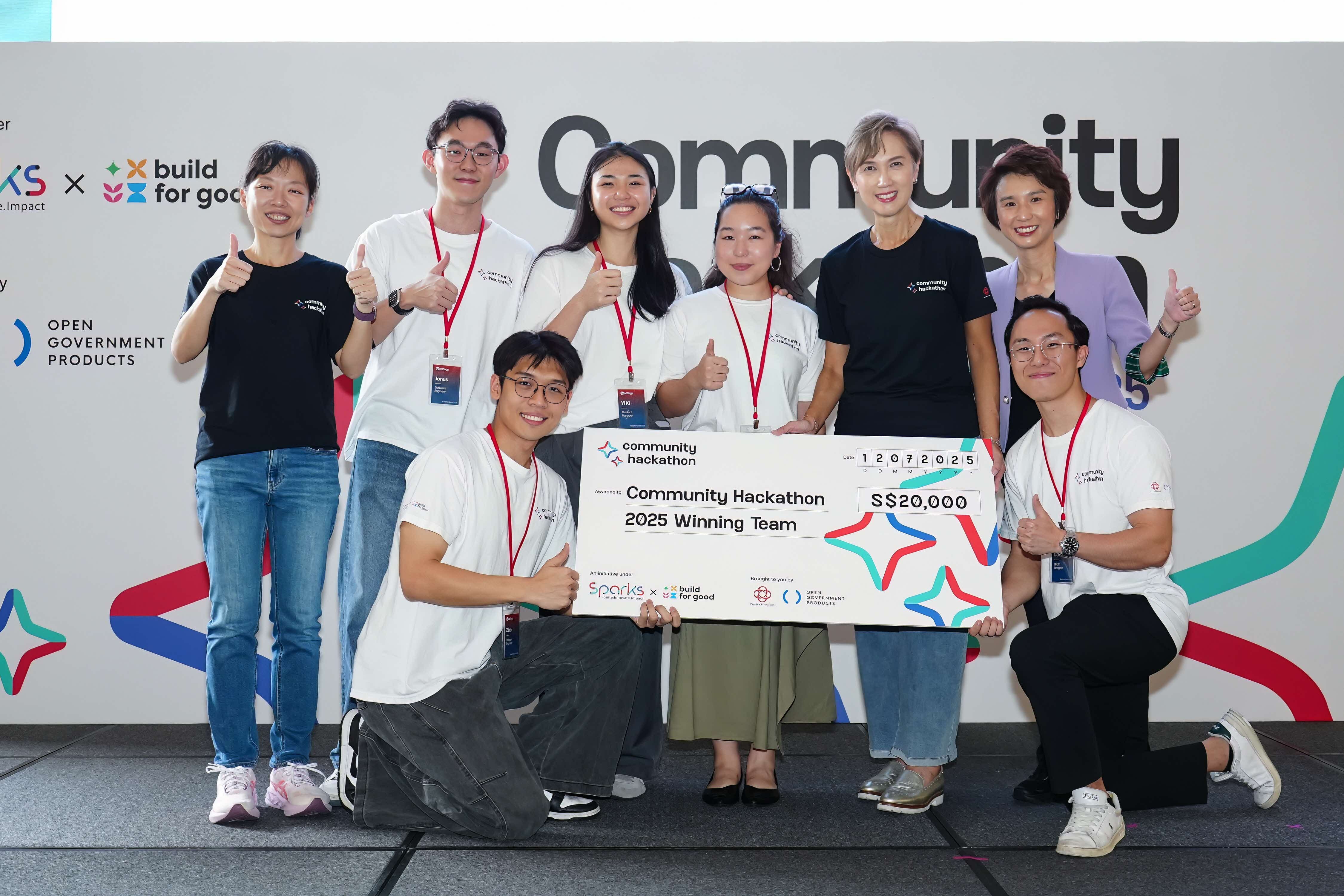 Eight people celebrating at a Community Hackathon event, with six standing in the back row giving thumbs up and two kneeling in front holding a large ceremonial cheque for S$20,000 made out to the "Community Hackathon 2025 Winning Team." Several participants wear red lanyards and matching white t-shirts. The backdrop shows "Community Hackathon" branding with logos for Sparks, Build for Good, and Open Government Products.