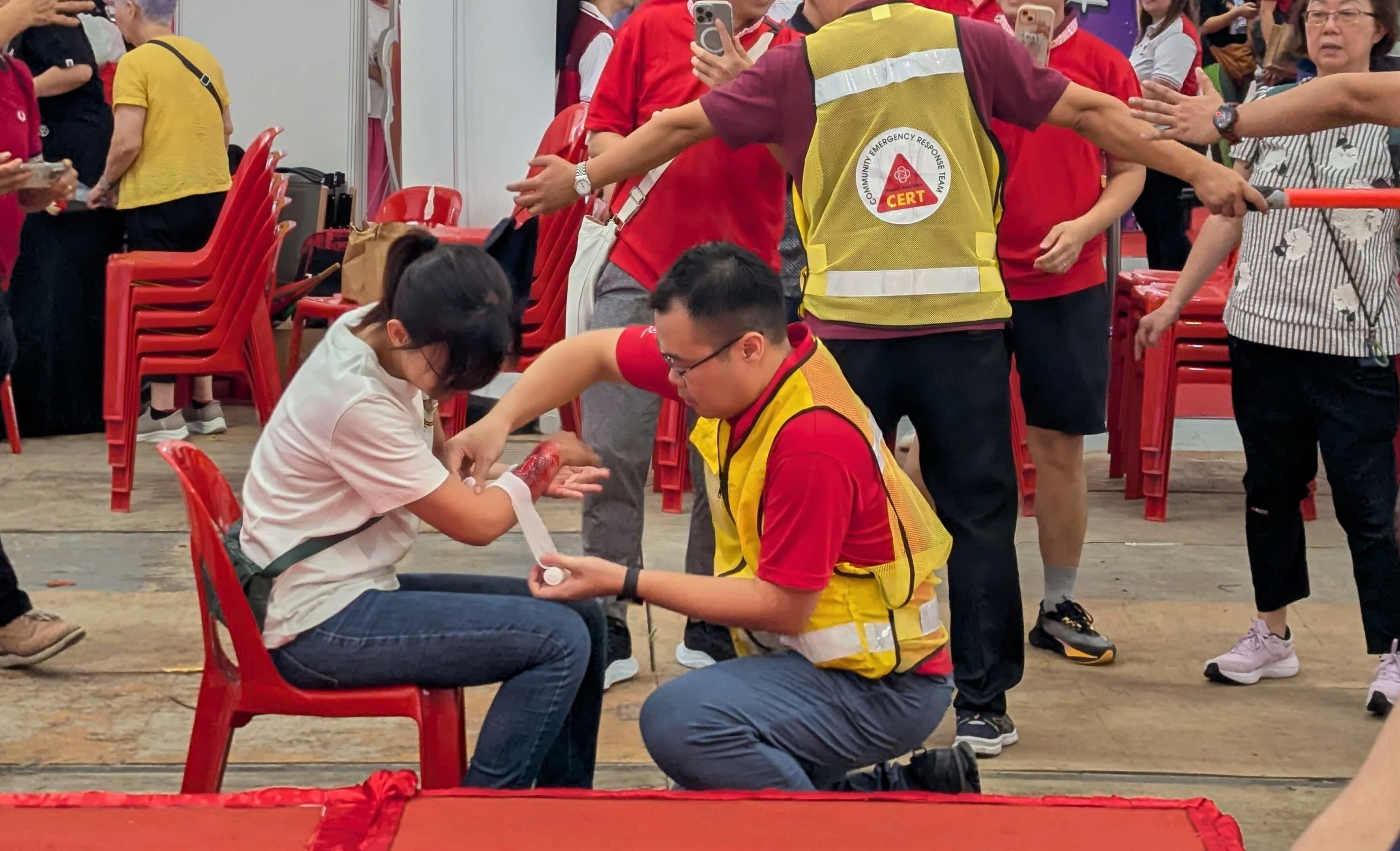 A person in a yellow vest applies a bandage to an individual's arm in a busy indoor setting.