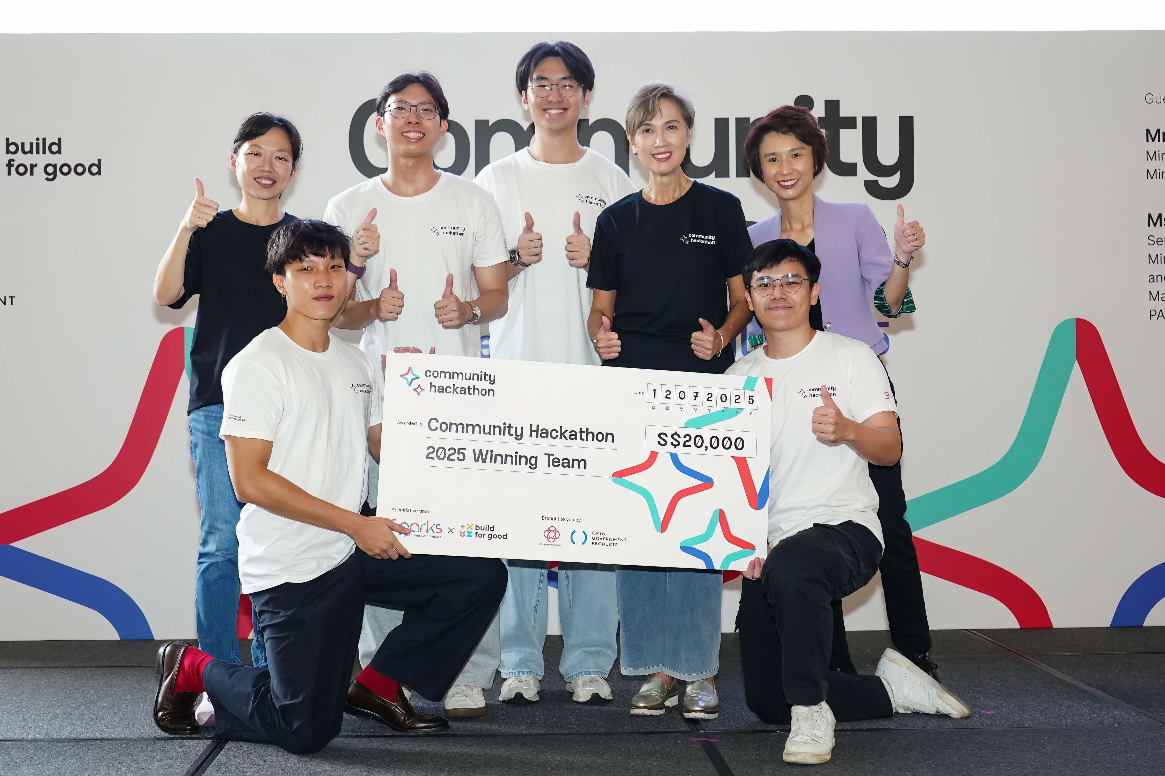 Seven people celebrating at a Community Hackathon event, with five standing in the back row giving thumbs up and two kneeling in front holding a large ceremonial cheque for S$20,000 made out to the "Community Hackathon 2025 Winning Team." Most participants wear white t-shirts with event branding. The backdrop shows "Community Hackathon" branding with "build for good" logo visible on the left side.