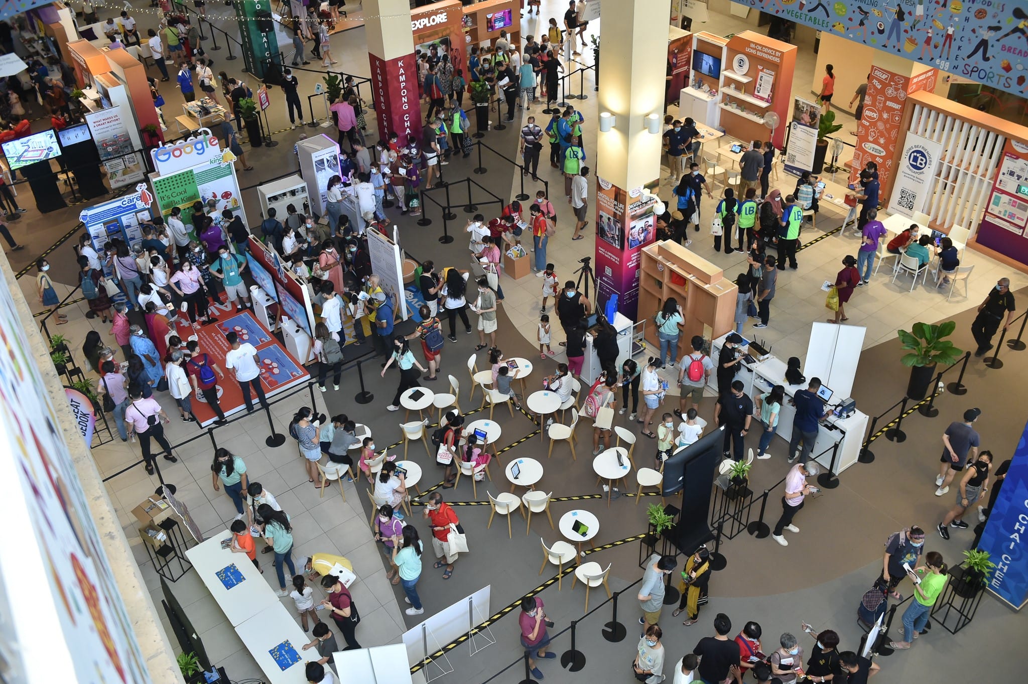 A busy indoor event with people interacting at various booths and tables, seen from a high vantage point.