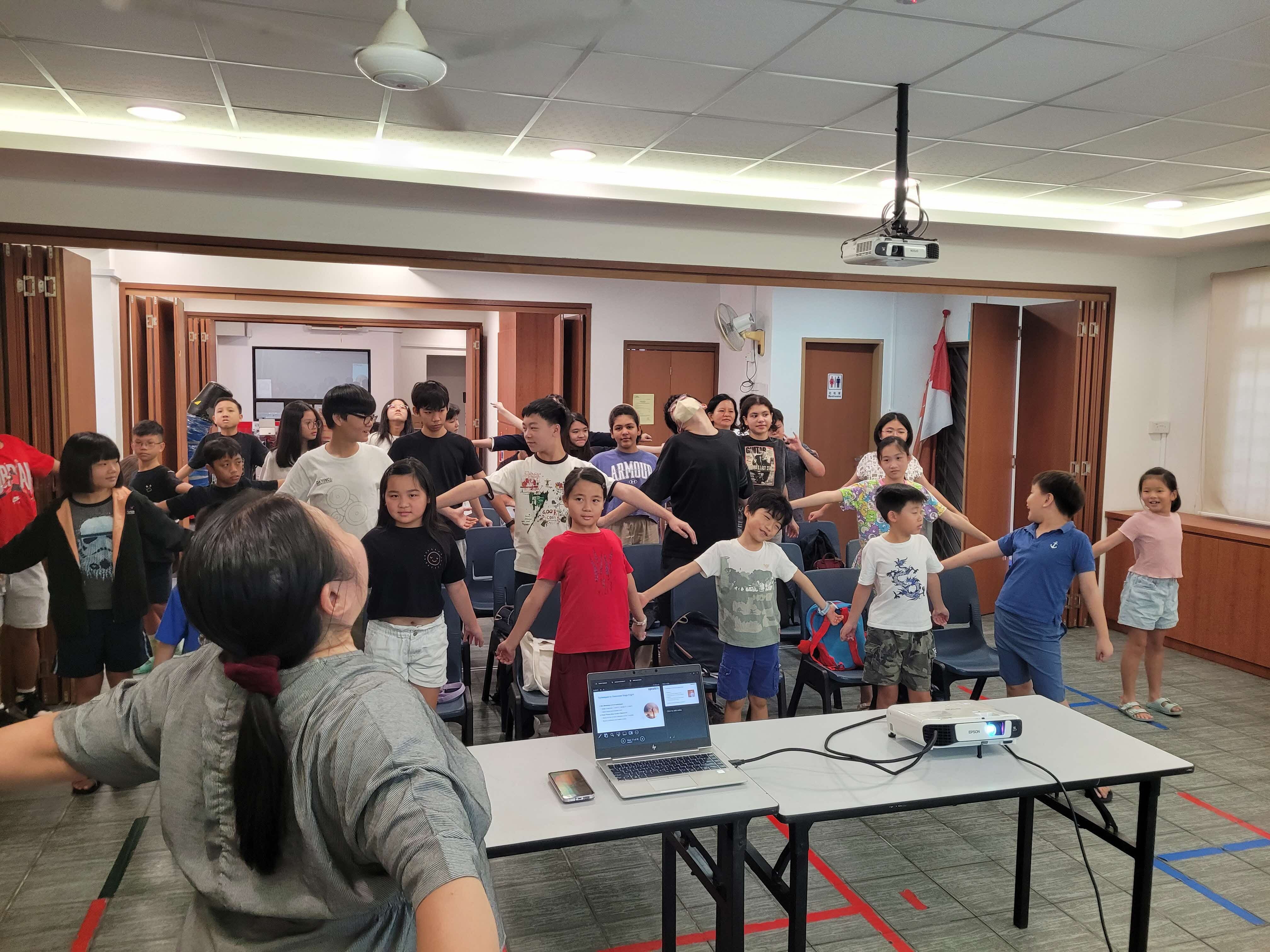 About 25-30 children and teenagers standing in a circle holding hands during a group activity in a modern conference room. An adult facilitator sits at a table with a laptop in the foreground.
