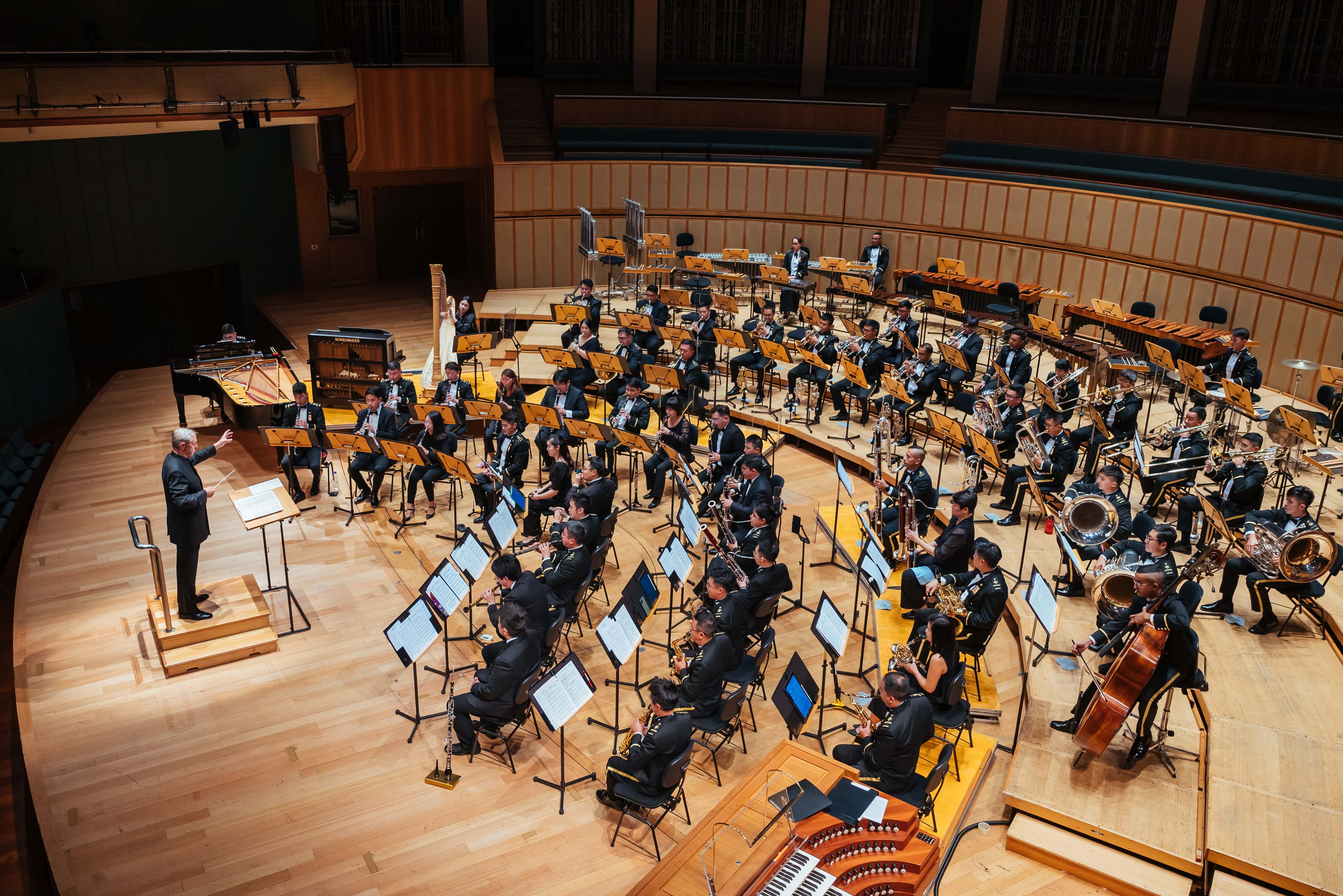 Military band in formal attire performs indoors under a conductor on stage.
