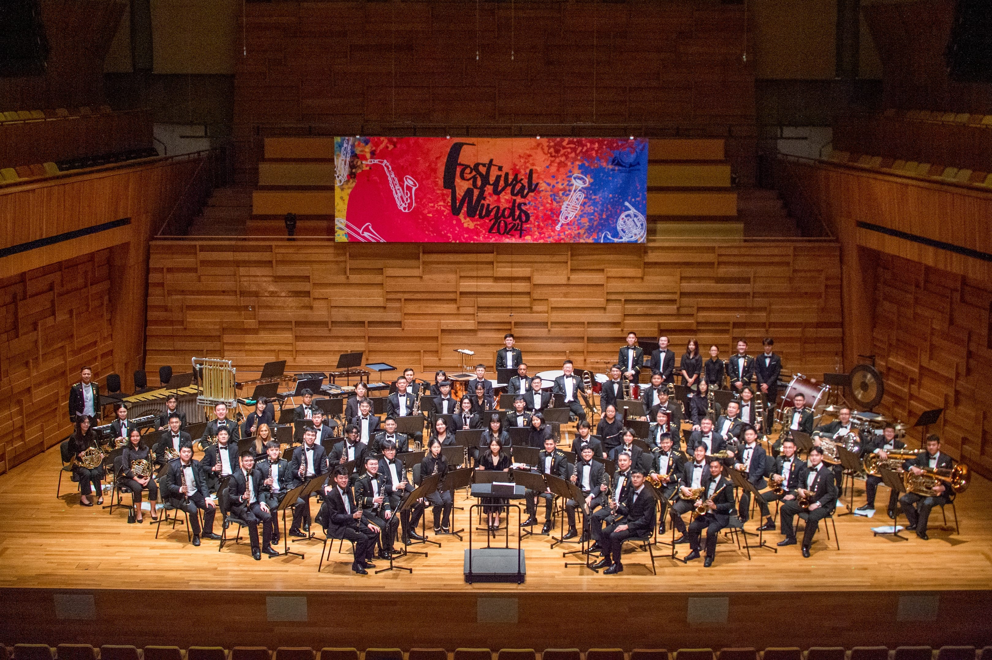Youth wind band posing in concert hall with instruments under “Festival Winds 2024” banner.
