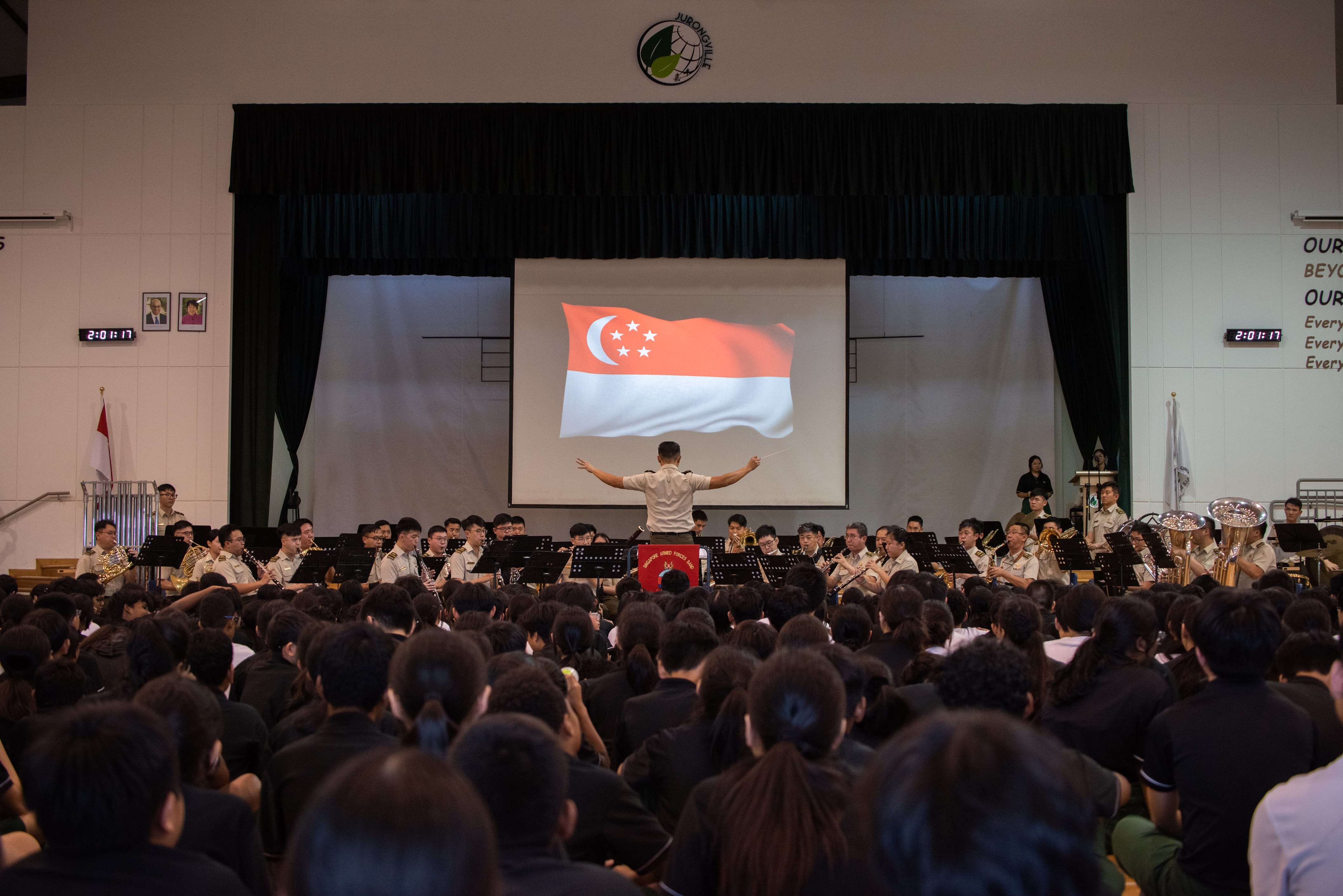 Band performing for school audience with conductor and Singapore flag projected on screen.