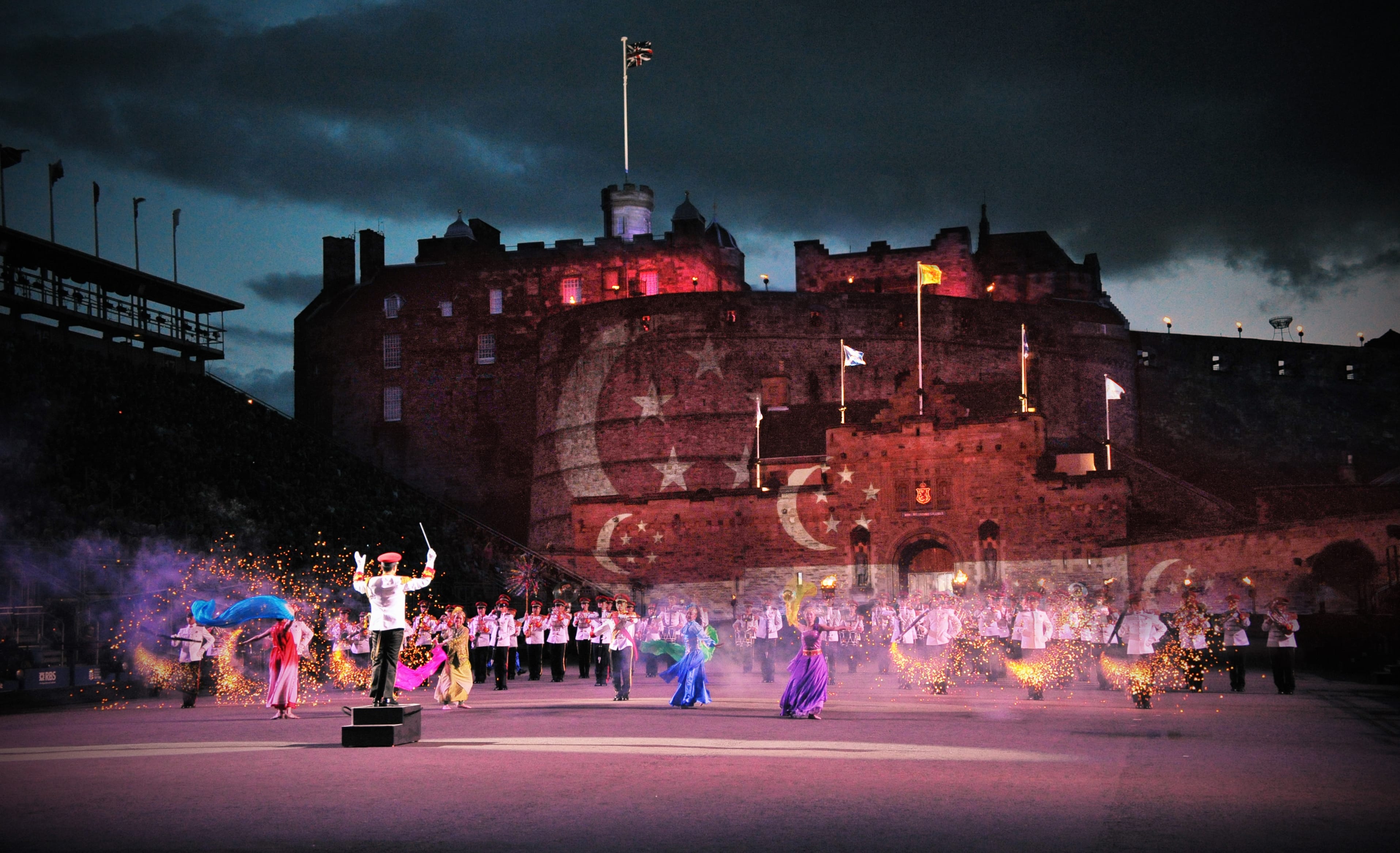 Military band and dancers perform at night in front of a lit-up castle with projected visuals.