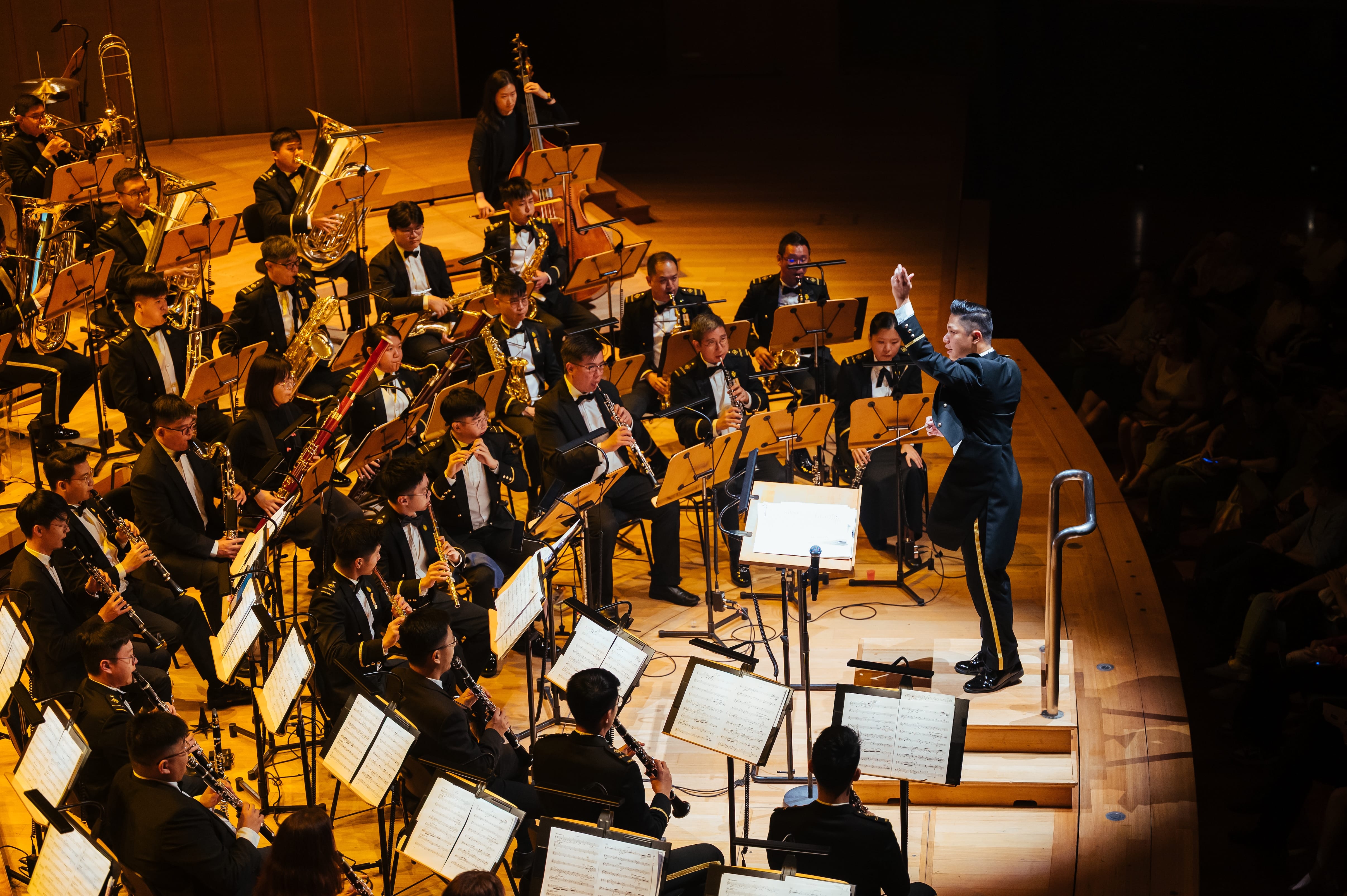 Conductor leading uniformed wind orchestra on concert stage with music stands and lighting.