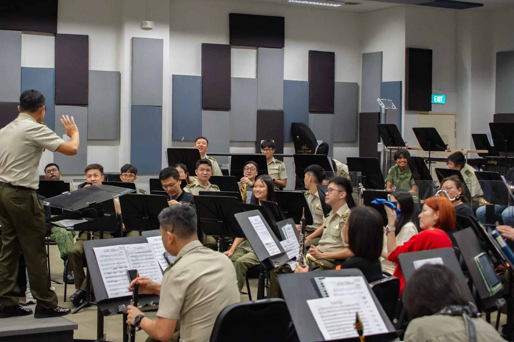 Conductor leading a military band, with musicians seated in a rehearsal room.
