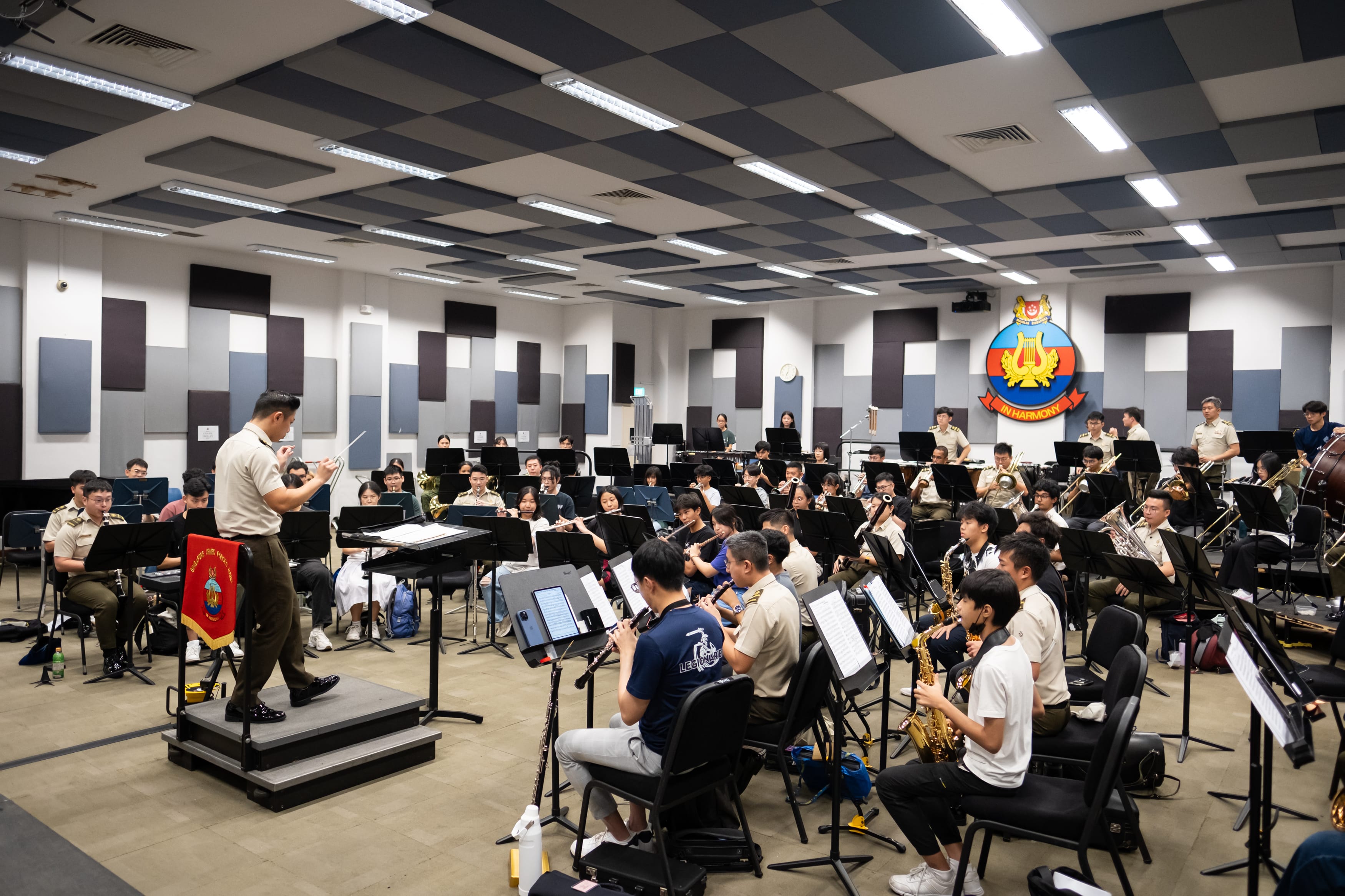 Large youth concert band rehearsing with conductor in acoustic room under "In Harmony" crest.