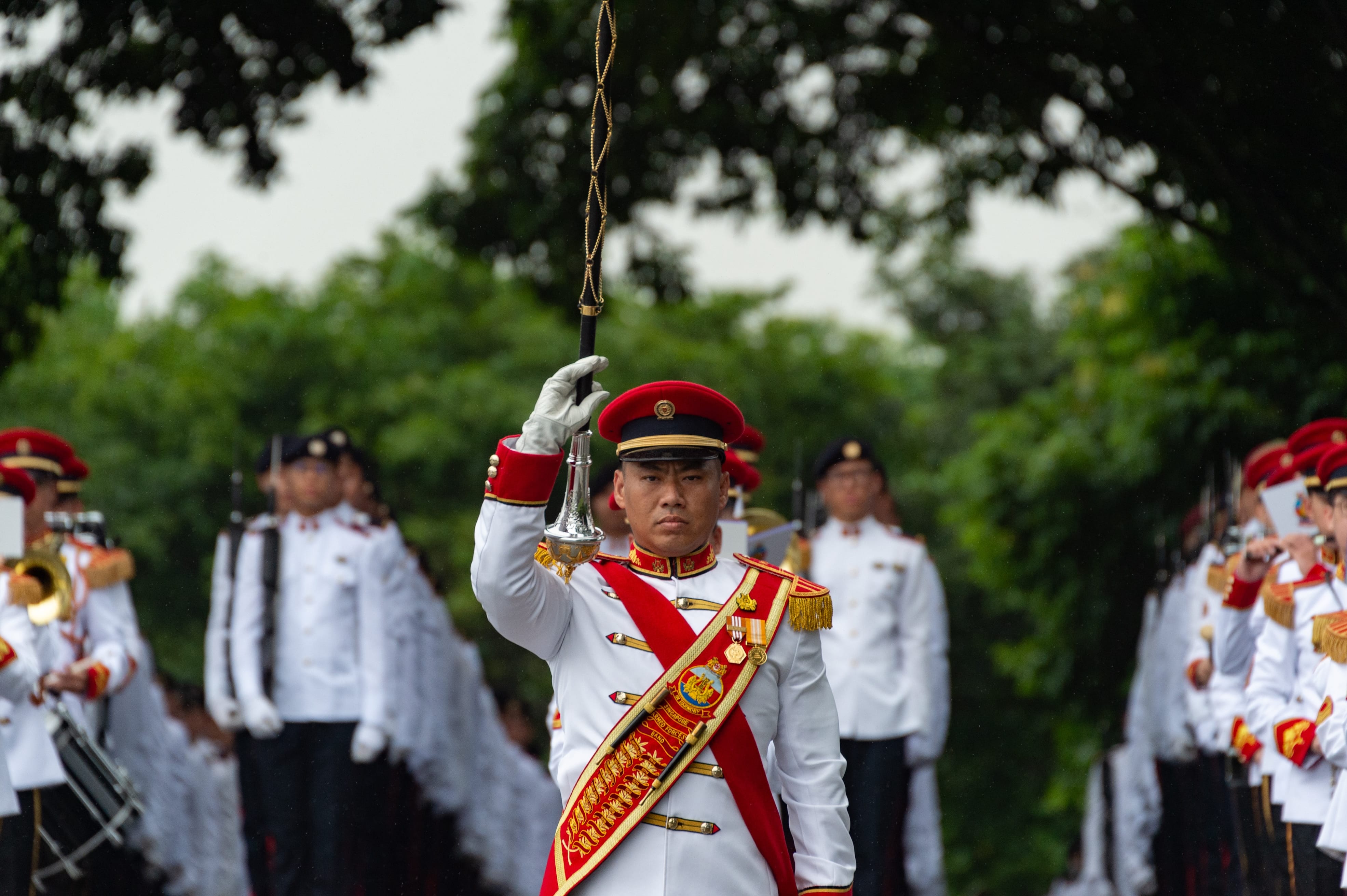 Drum major in ceremonial uniform leading a military band with mace raised during parade.