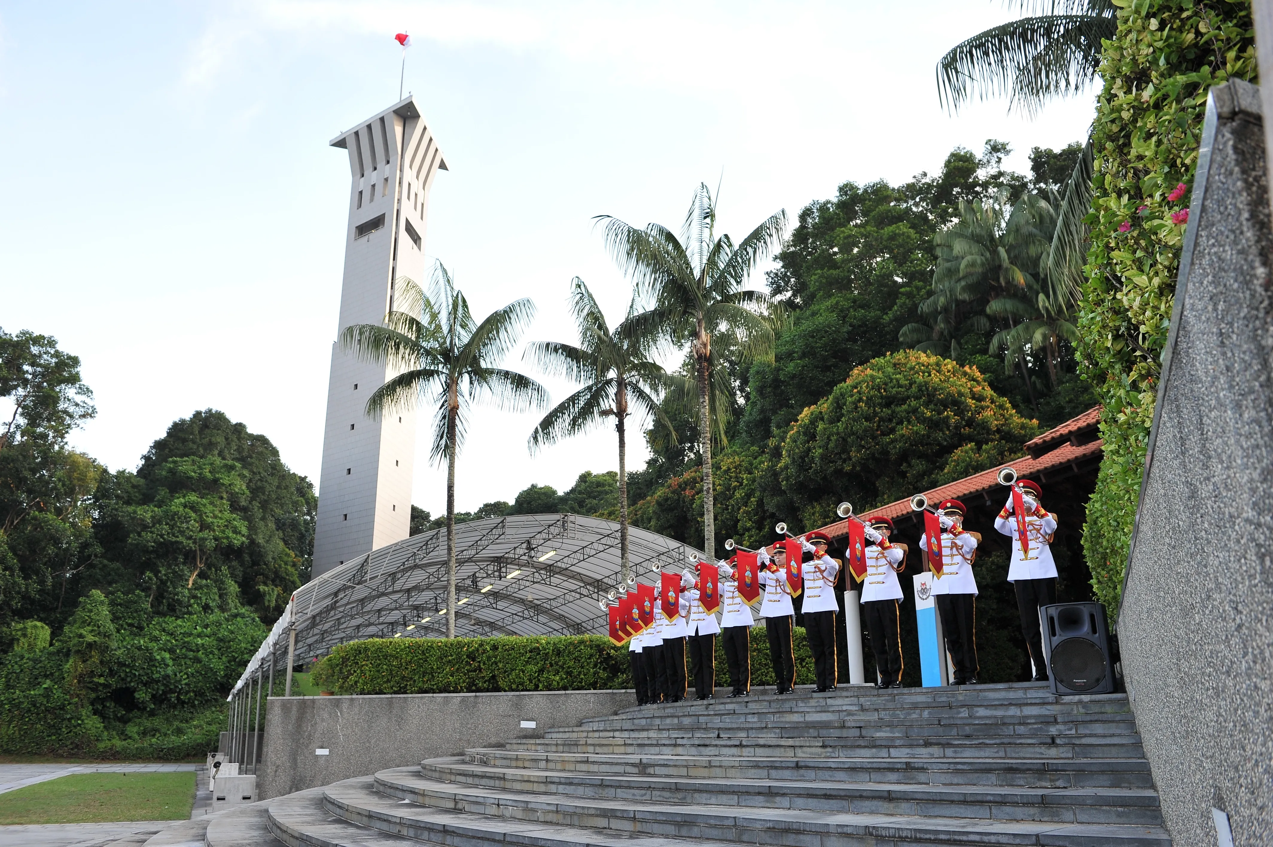 Uniformed military band playing bugles at steps of war memorial, with SAFTI tower behind.