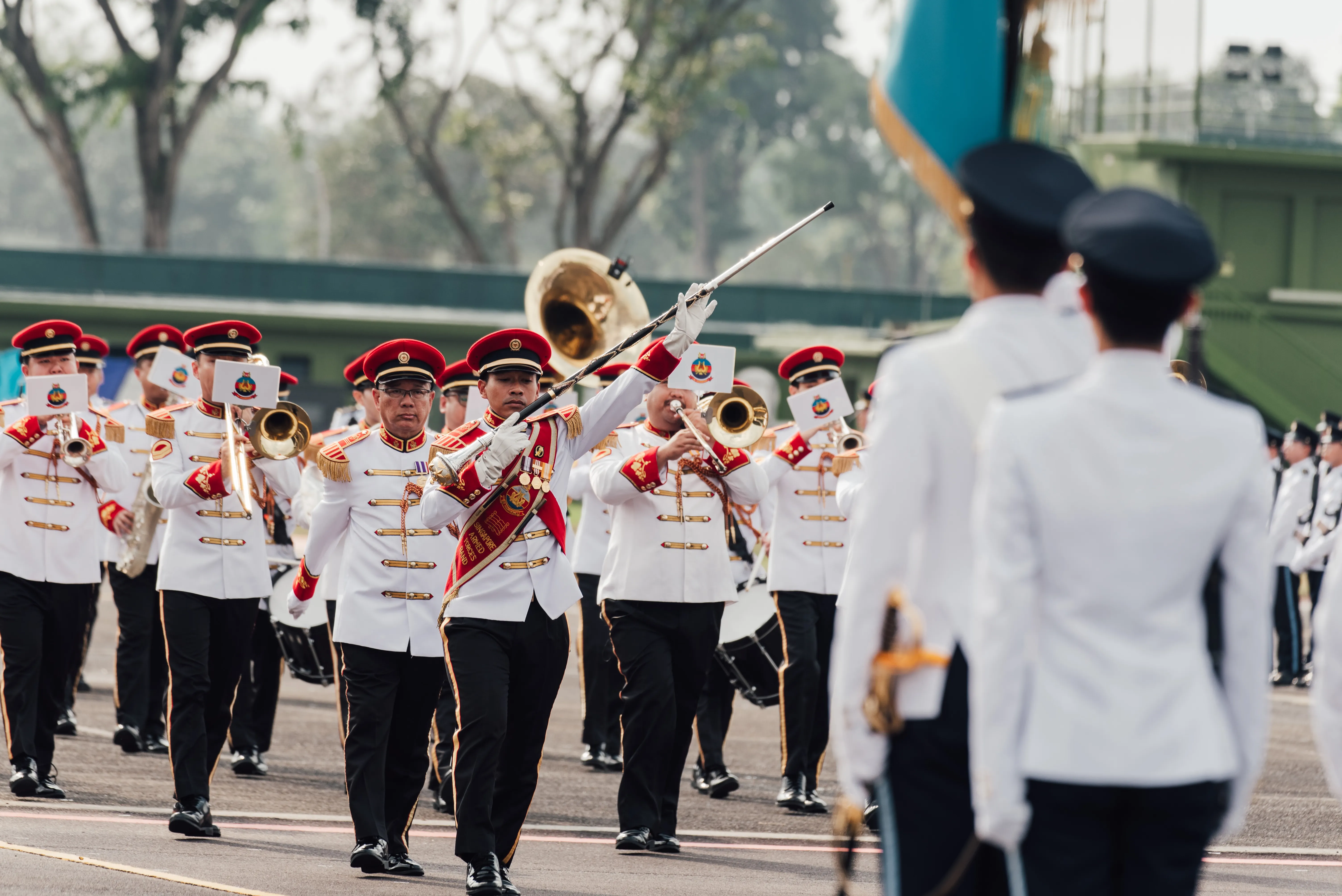 Military band in white ceremonial uniforms marches with instruments and conductor leading in front.