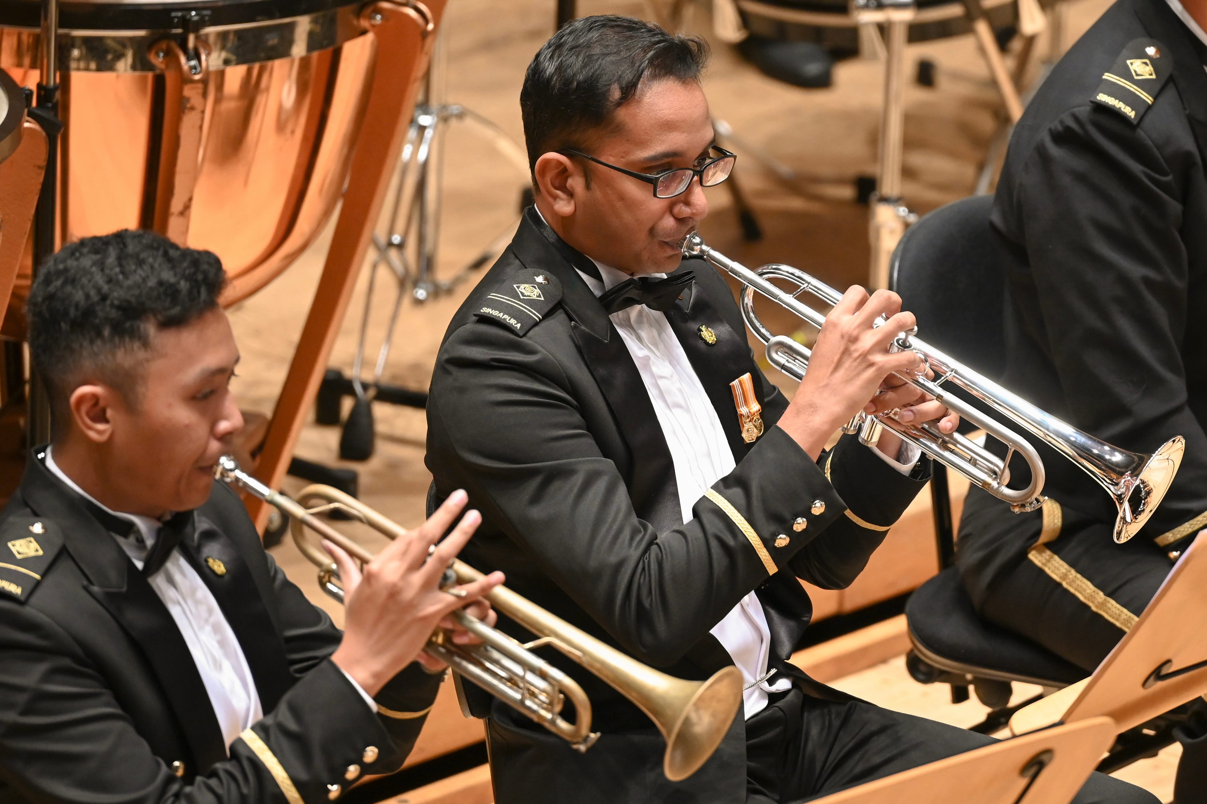 Brass musicians in formal band uniforms performing with trumpets and trombone on concert stage.