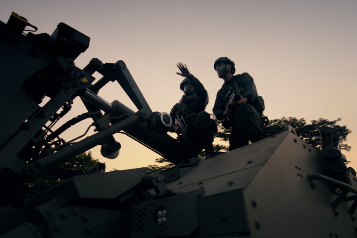 Two soldiers gesture on top of large combat vehicle during dusk deployment.