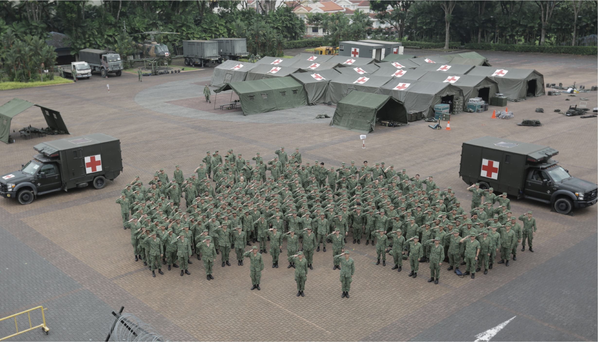 Soldiers saluting in formation with field hospital tents and medical vehicles in the background.