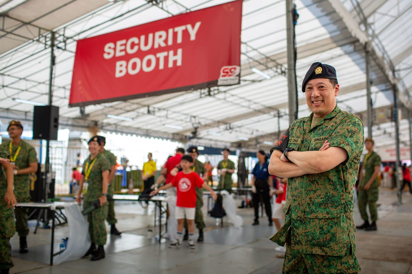 Uniformed officer at NDP security booth smiling while crowd passes through.