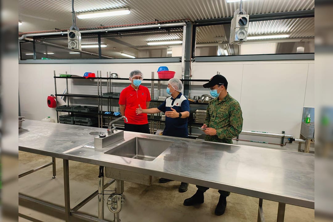 Officers and a soldier tour a camp kitchen wearing hairnets and masks.