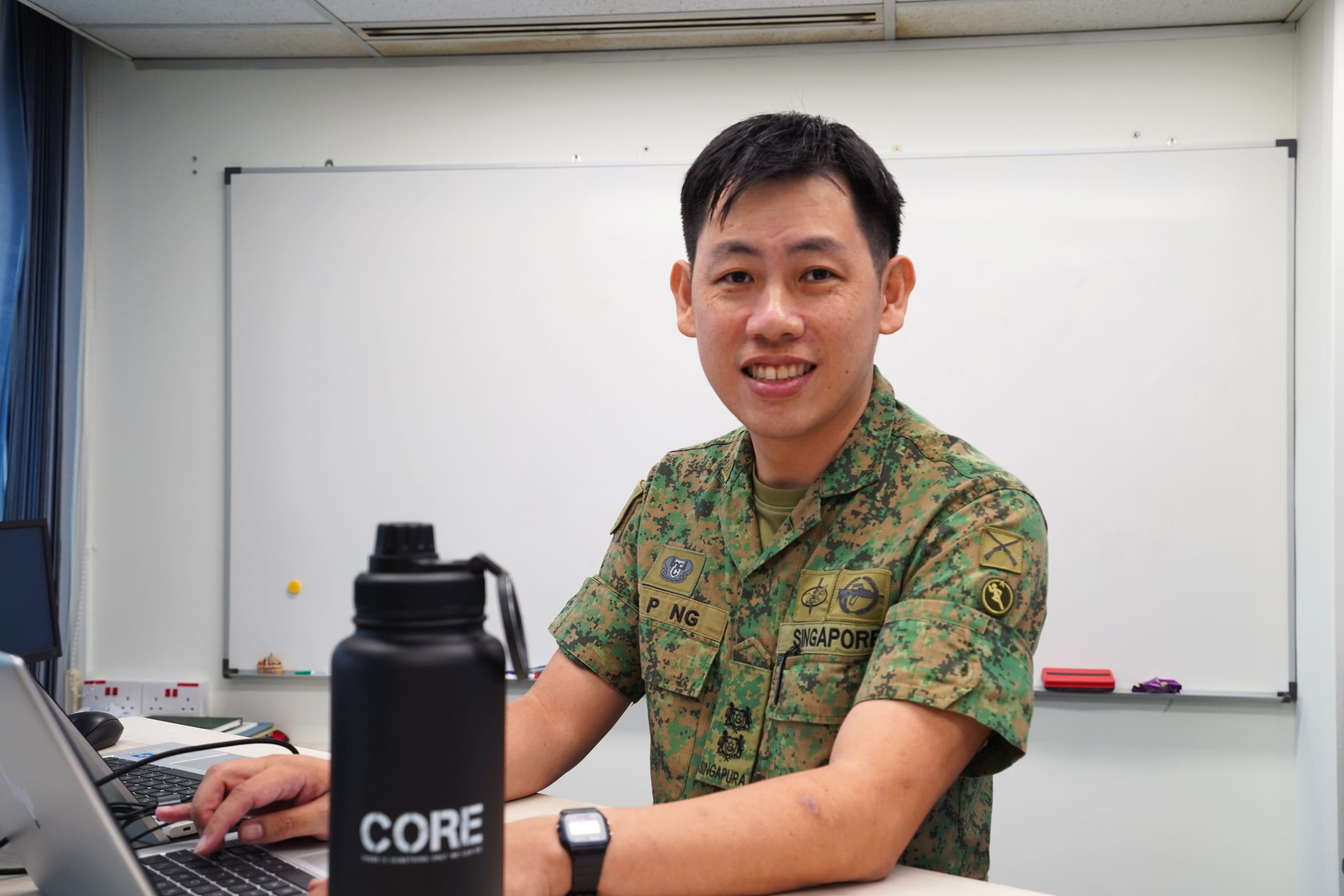 LTC Ng Tze Peng smiling at his desk with a laptop and CORE bottle in front.
