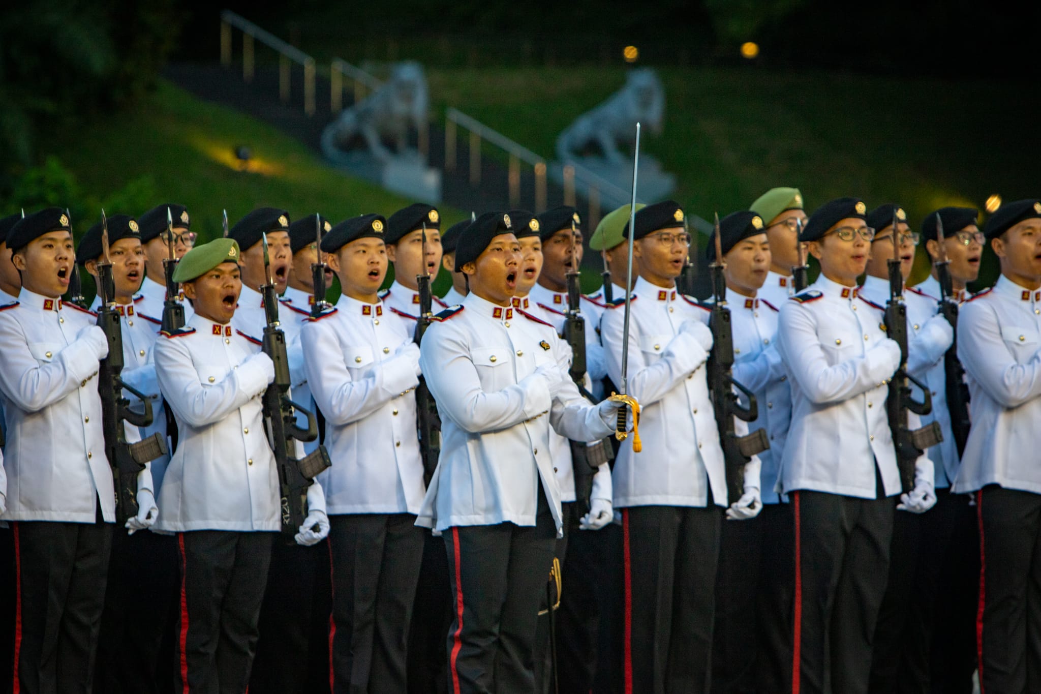 A formation of uniformed soldiers in white ceremonial dress, rifles in hand, and right fists over their hearts, during a formal parade.