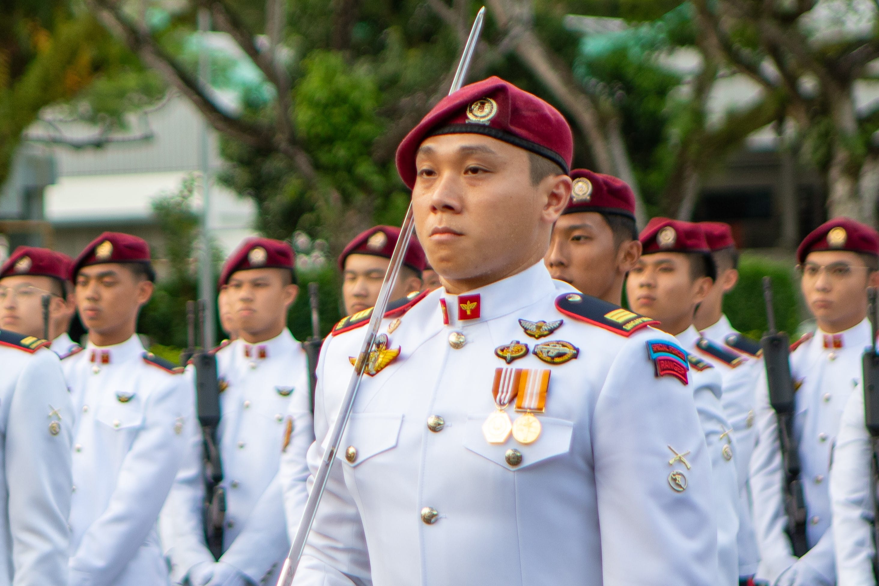 A ceremonial parade featuring a solemn Warrant Officer in white military dress uniform with medals, leading a group of soldiers in maroon berets.