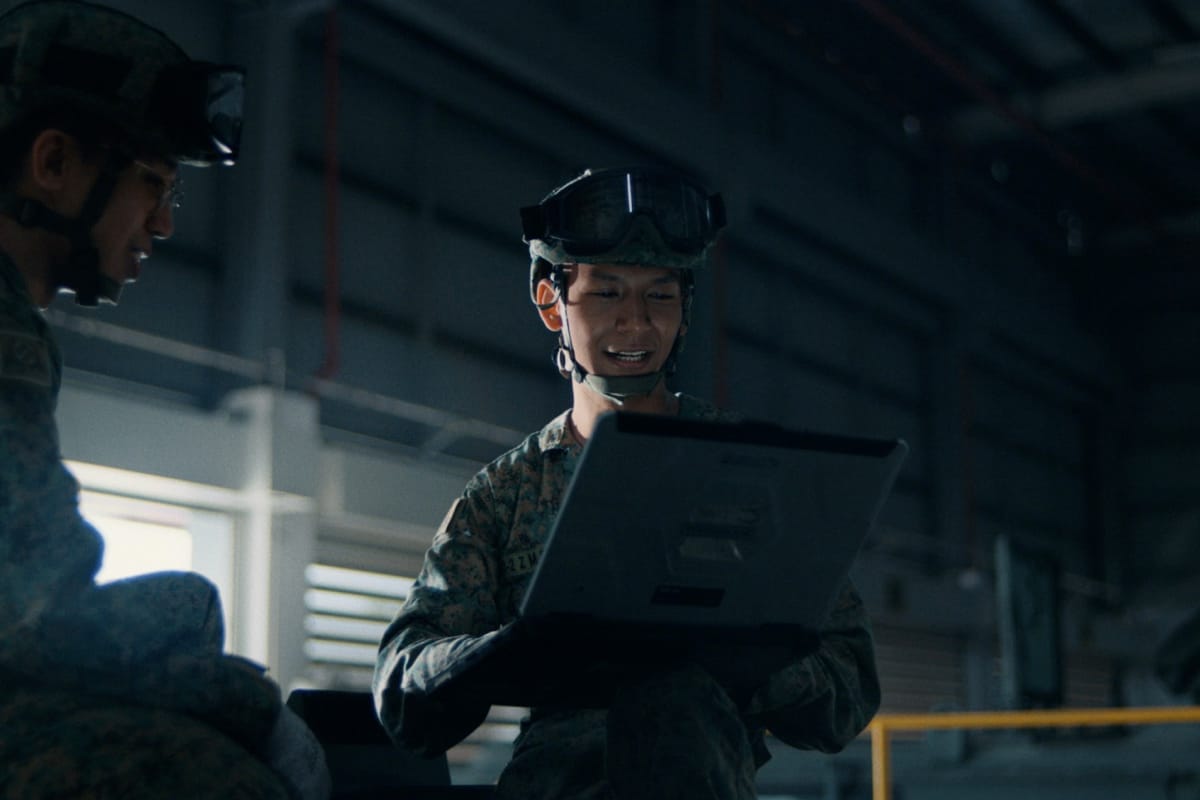 Soldiers review data on rugged laptop in military hangar under soft lighting.
