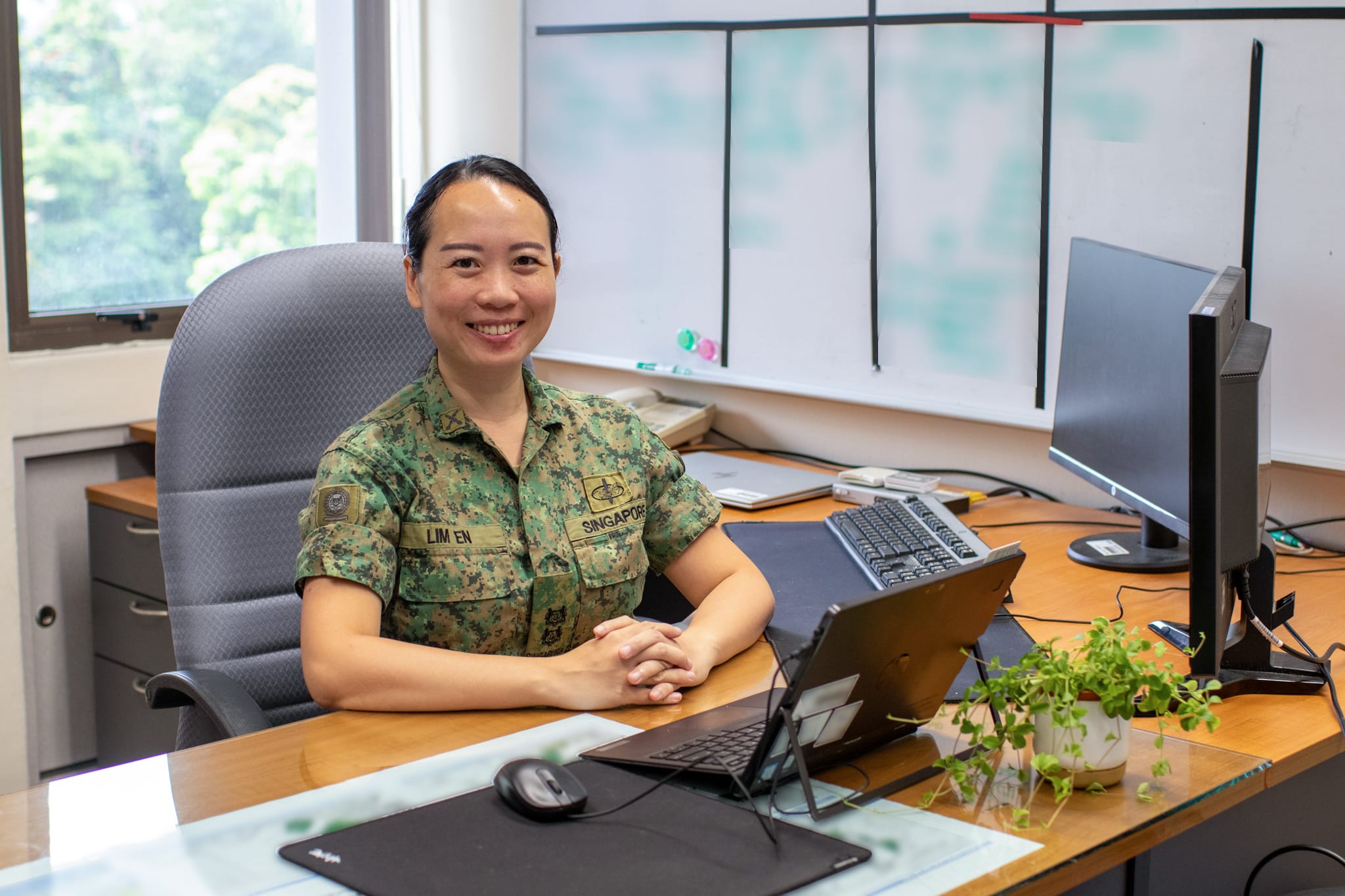 LTC Lim En at her desk in uniform, smiling in front of a whiteboard and laptop.