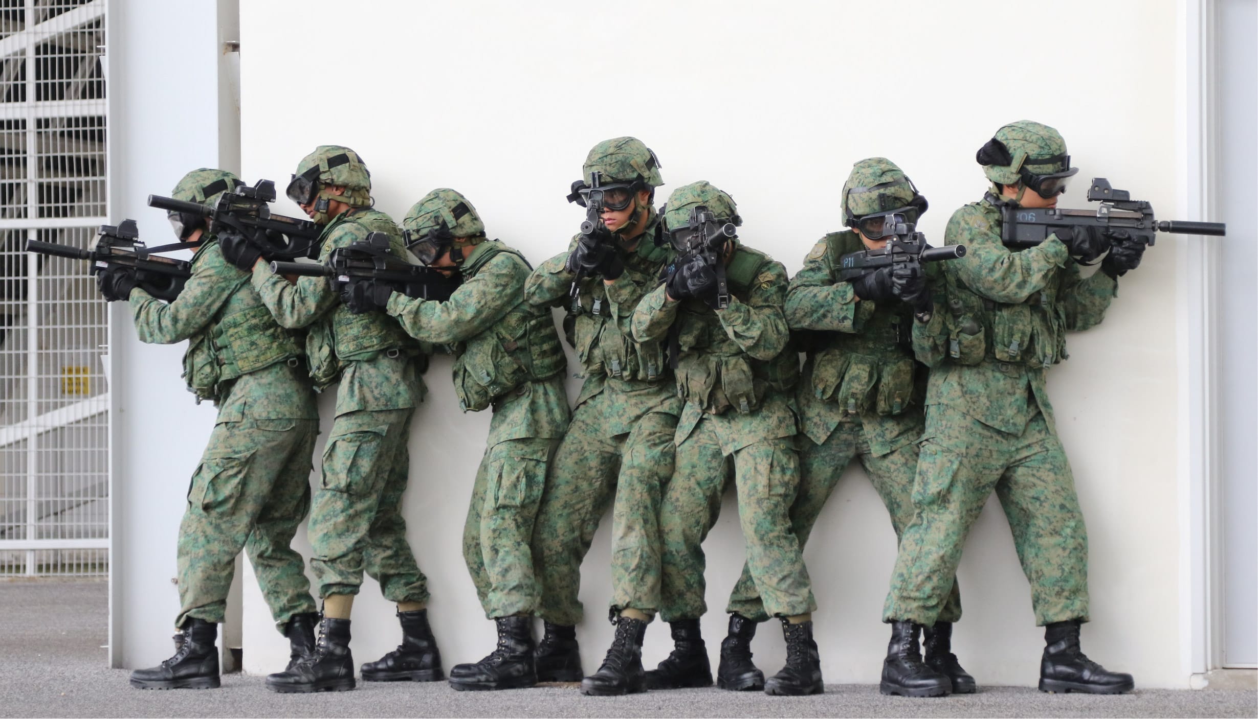 Soldiers in combat gear taking cover and aiming rifles during an urban training exercise.