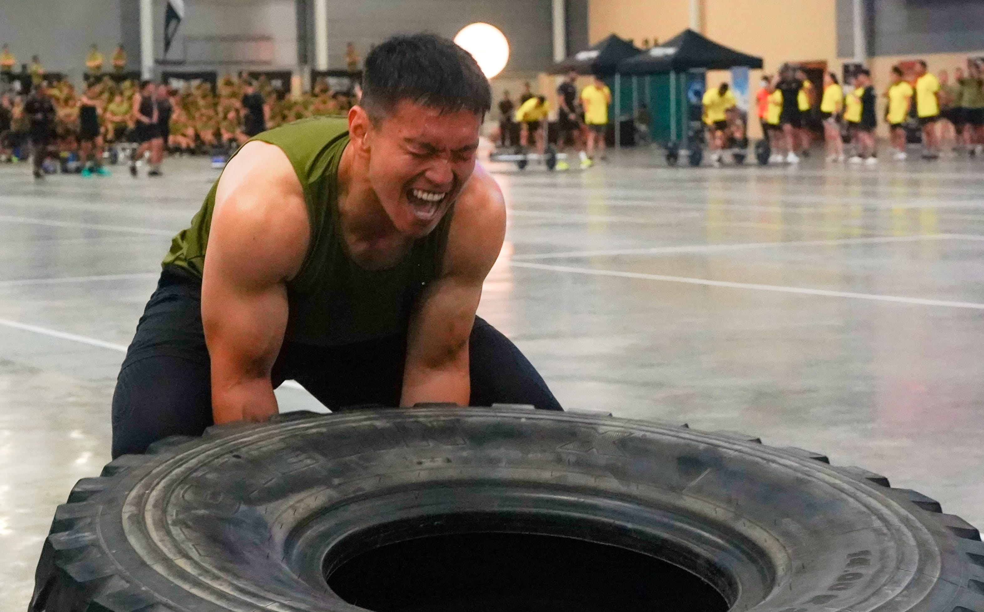 A male soldier straining to lift a large tire in an indoor fitness challenge, showcasing physical strength and determination.
