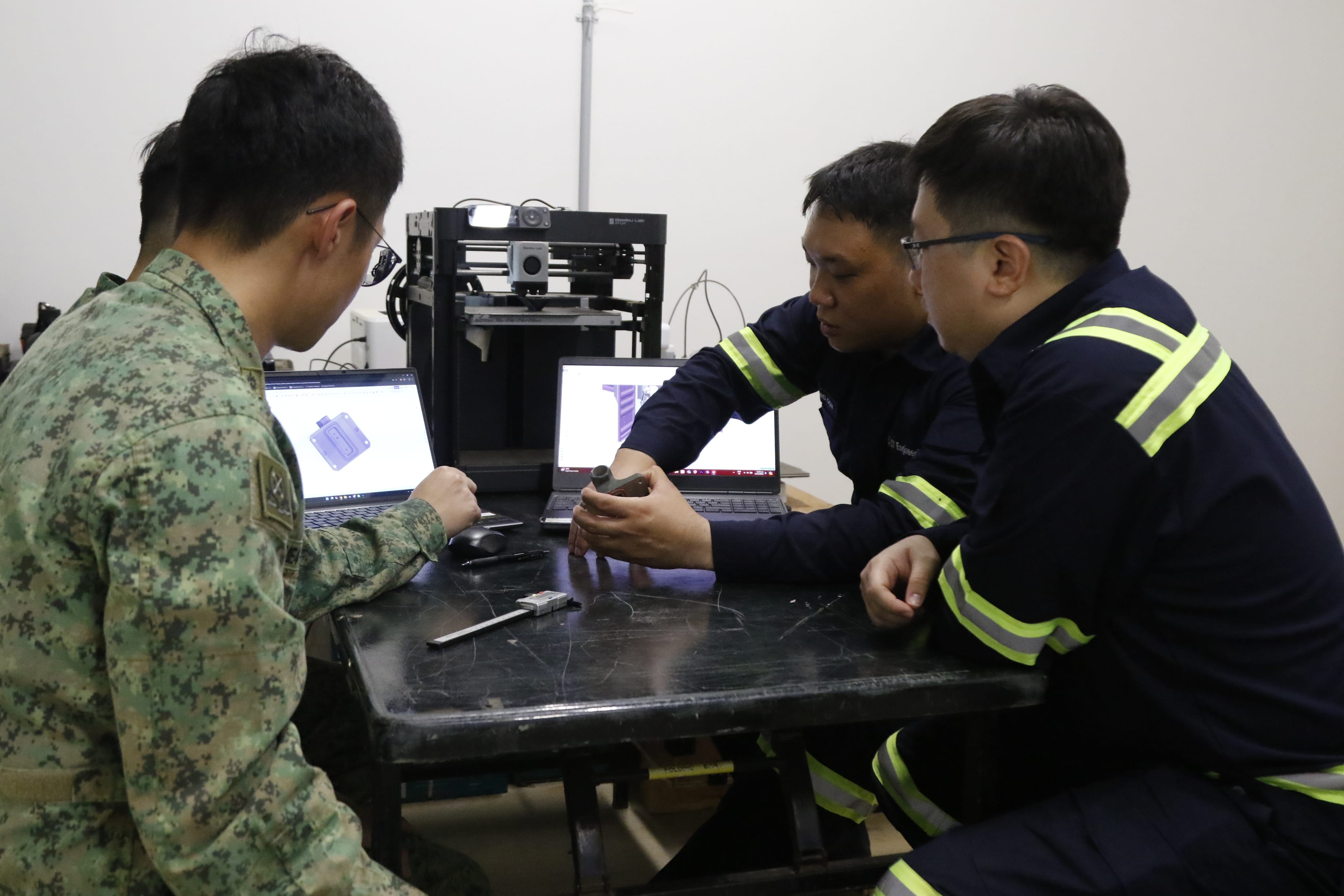 Soldier and engineers review 3D printed part using laptops and design tools.