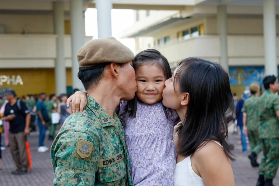 A soldier kisses his daughter while his wife kisses her cheek, smiling at an event.