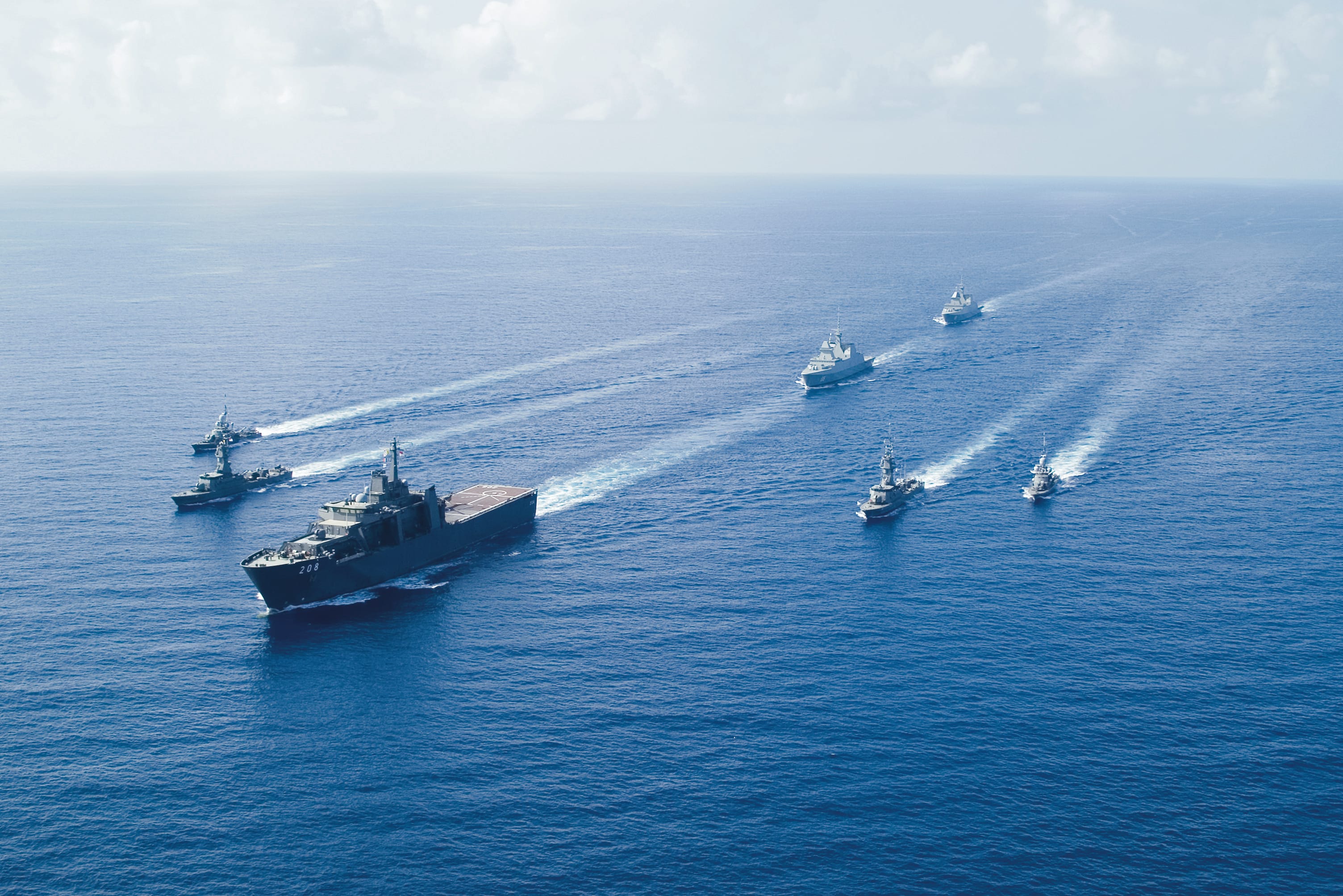 A formation of naval ships sails in parallel across deep blue waters during a coordinated exercise.