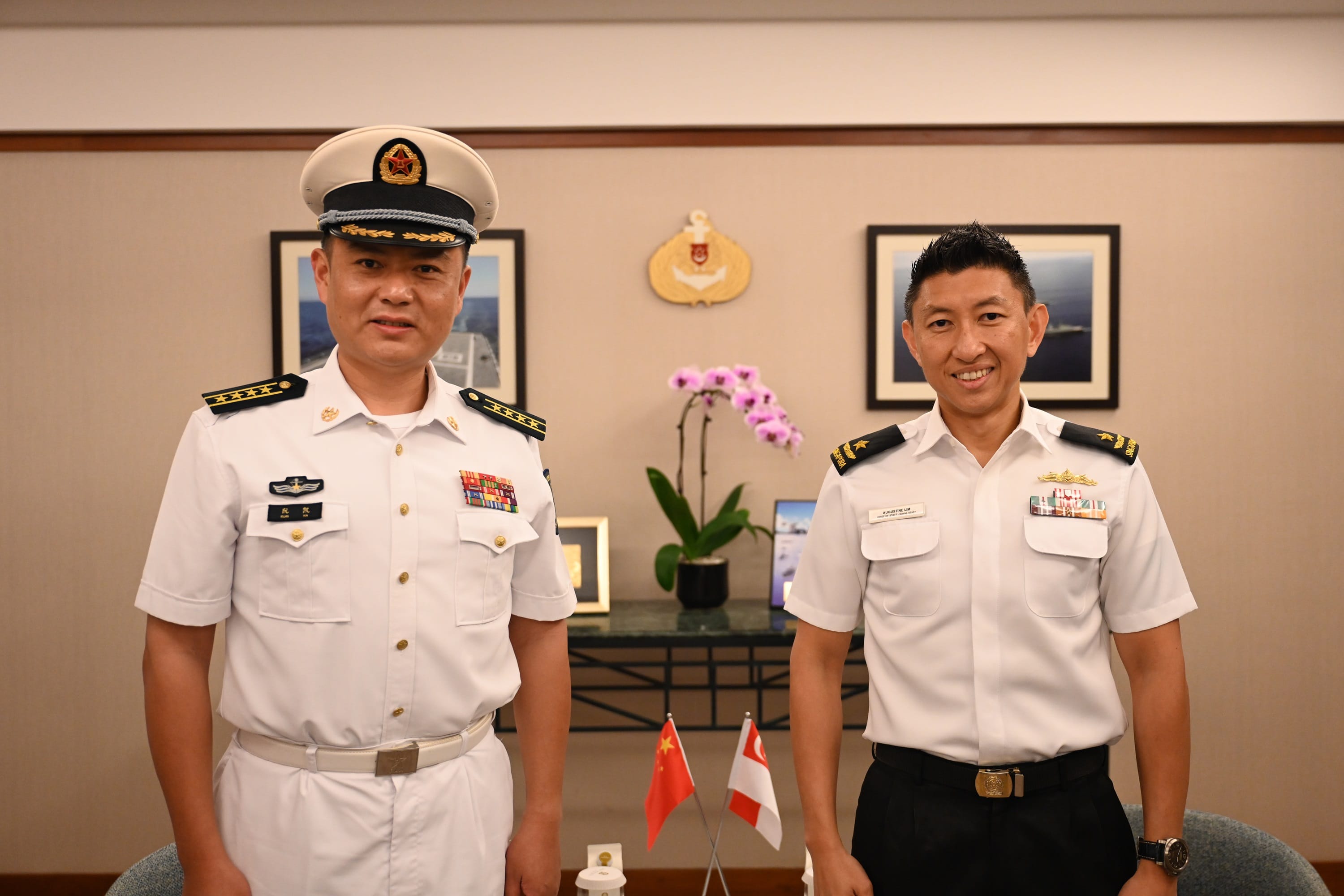 Two naval officers in uniform stand in a room with Chinese & Singaporean flags.