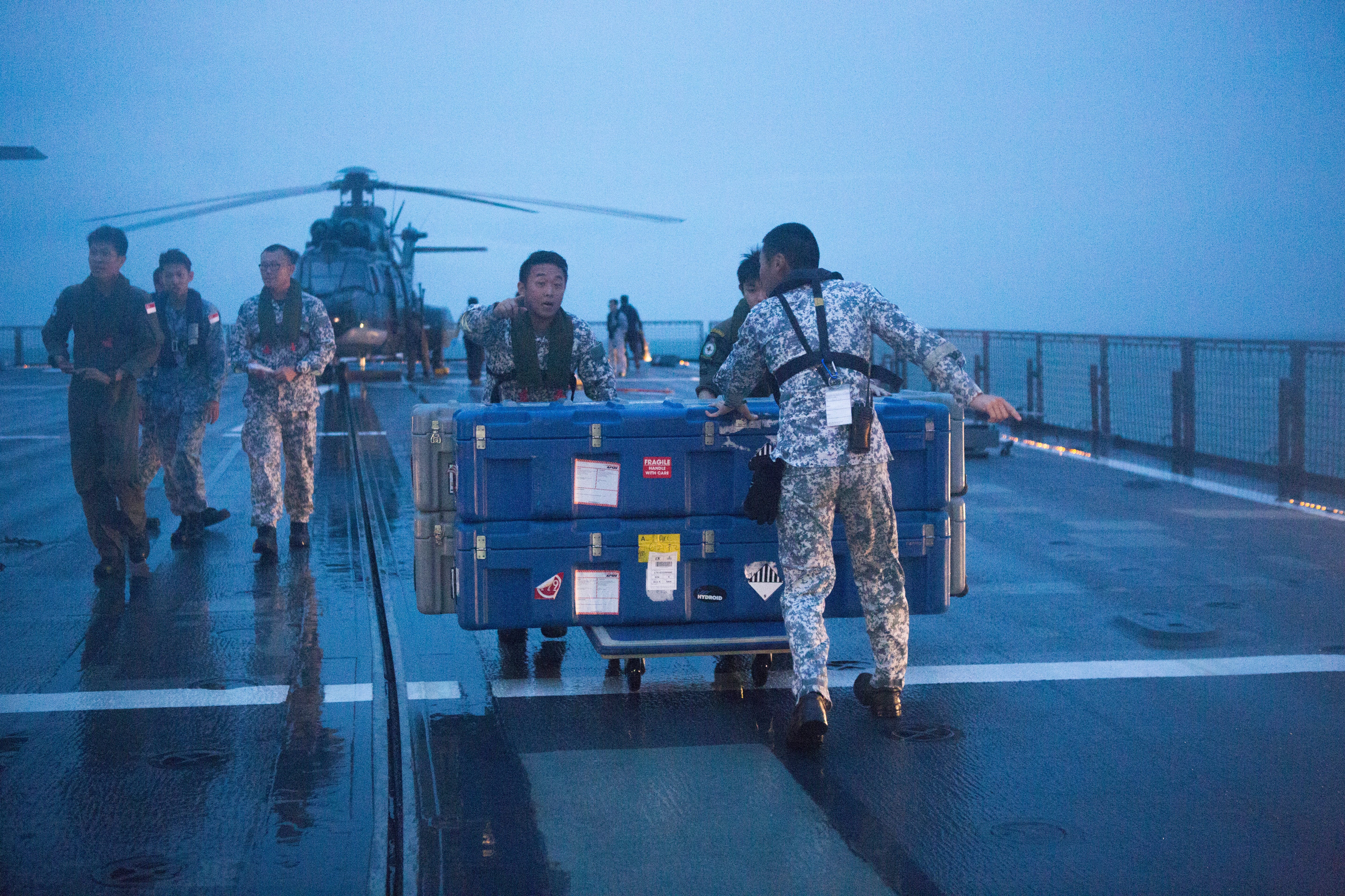 Uniformed personnel push large blue containers on a wet ship deck near a helicopter at dusk.