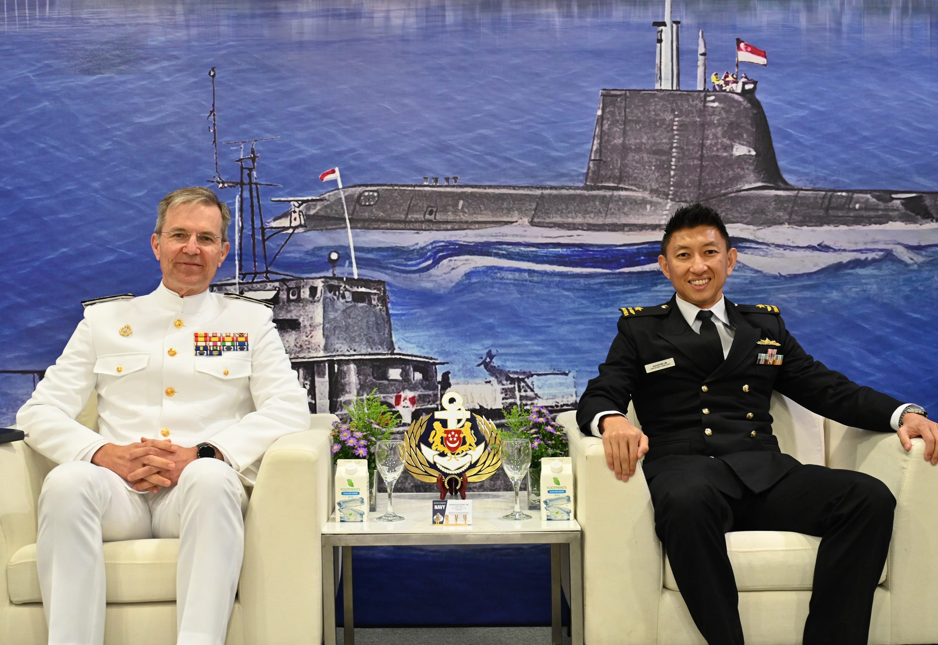 Two naval officers from Spain and Singapore seated in front of an illustrated maritime backdrop.