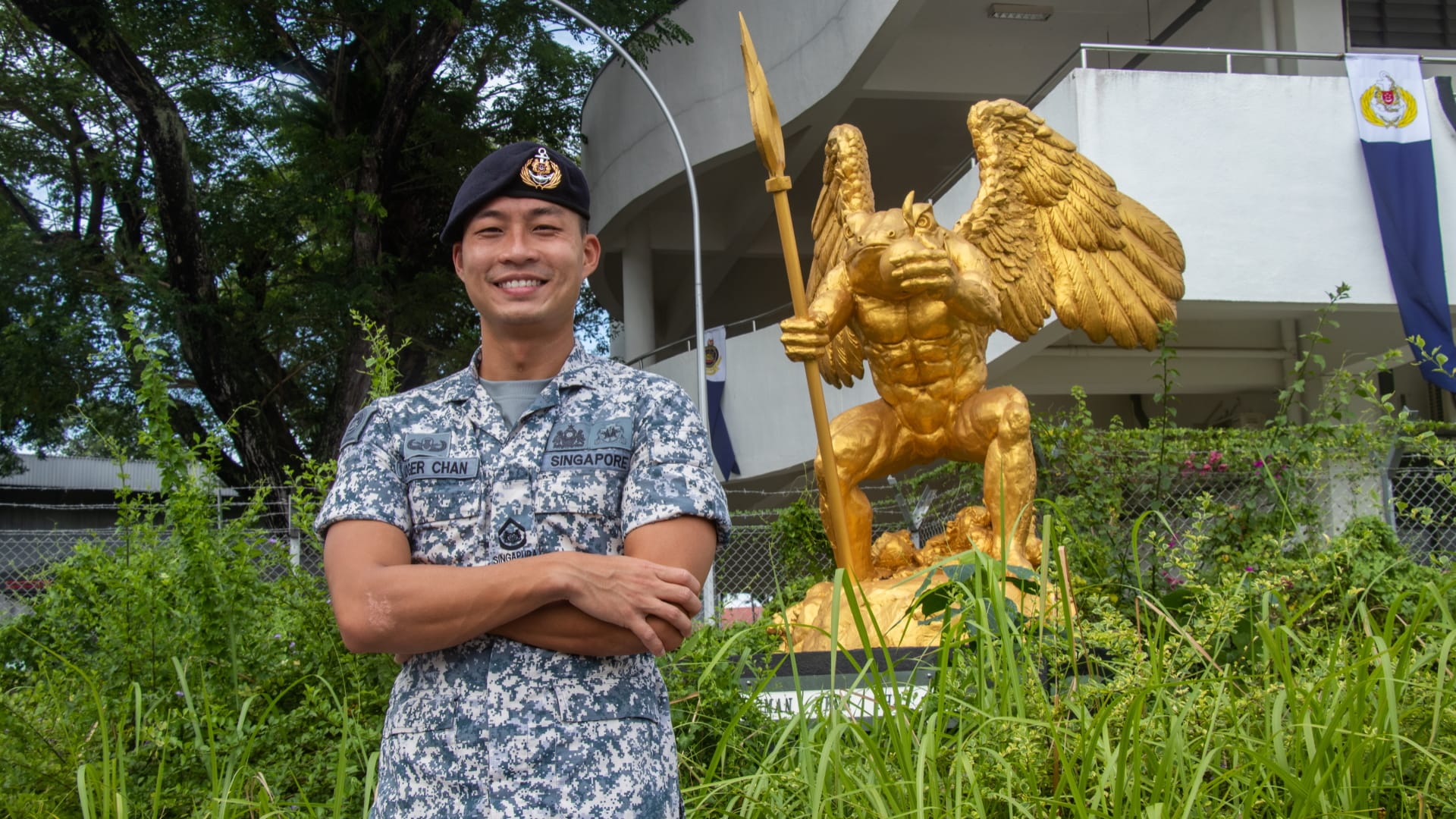 2WO Roger Chan stands smiling in Navy uniform beside a golden griffin statue holding a spear.