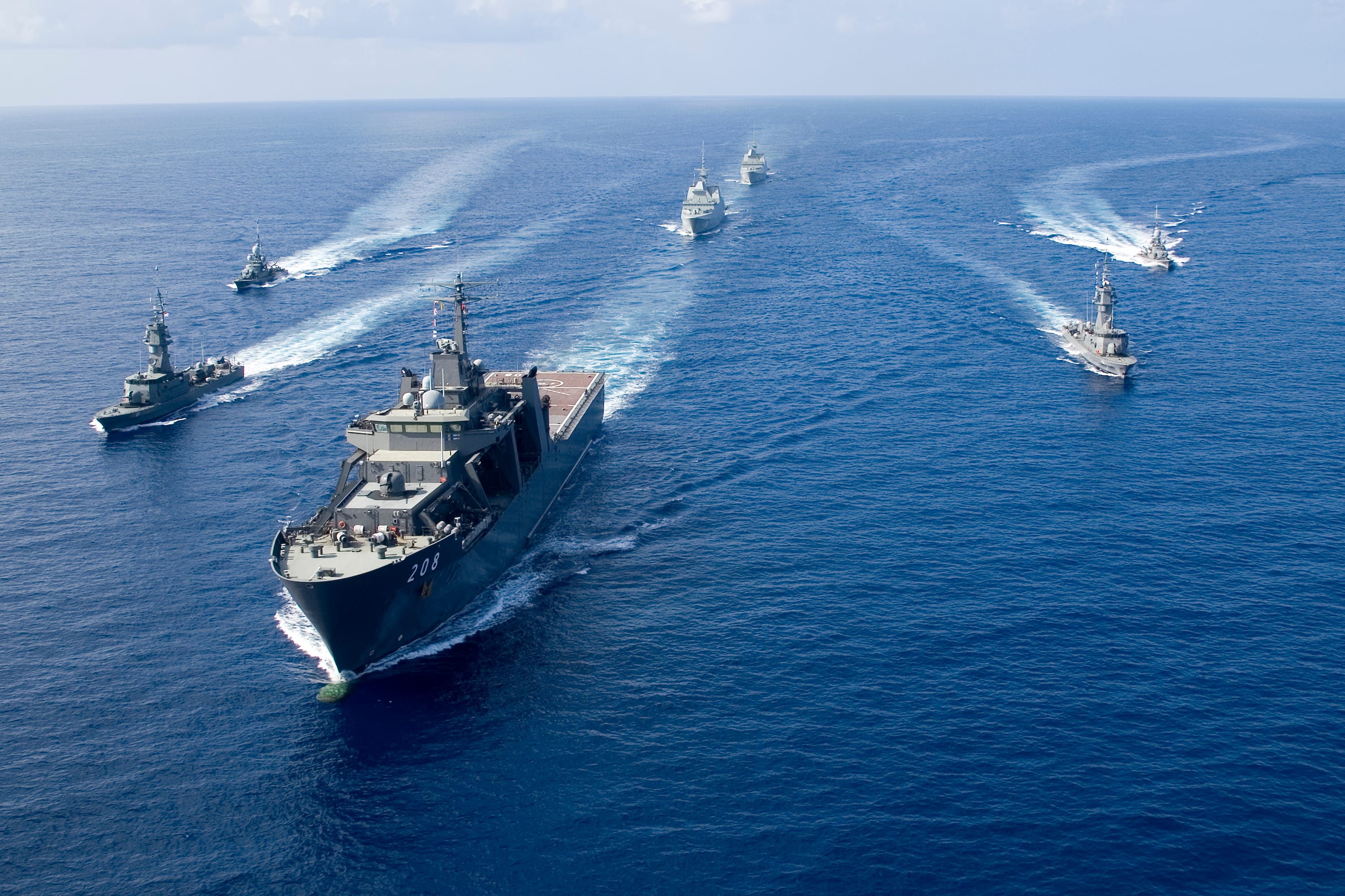 A fleet of naval ships sails in tight formation, creating white wakes behind them on open ocean waters.