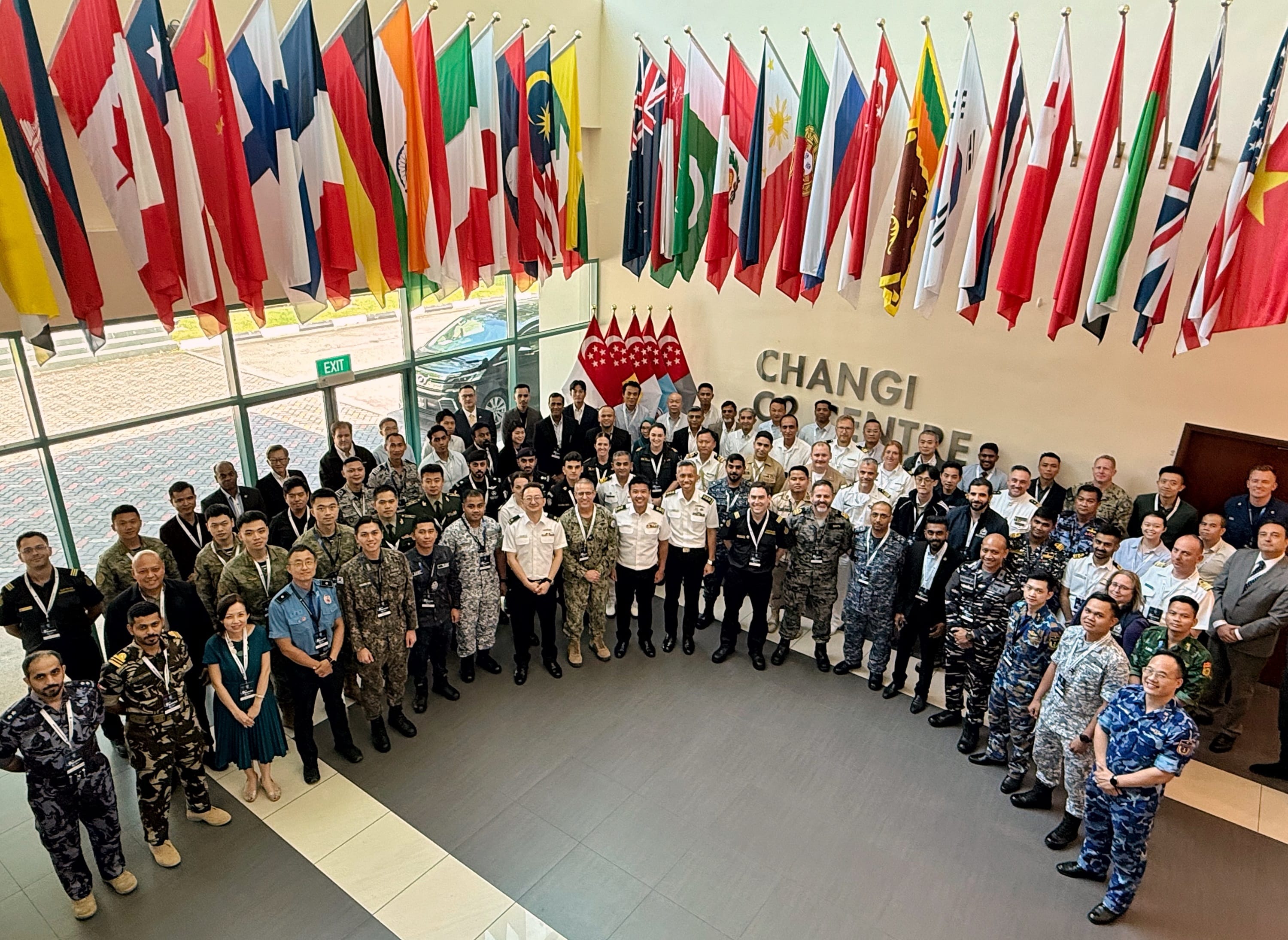 Large group of uniformed personnel posing under international flags at Changi Centre.