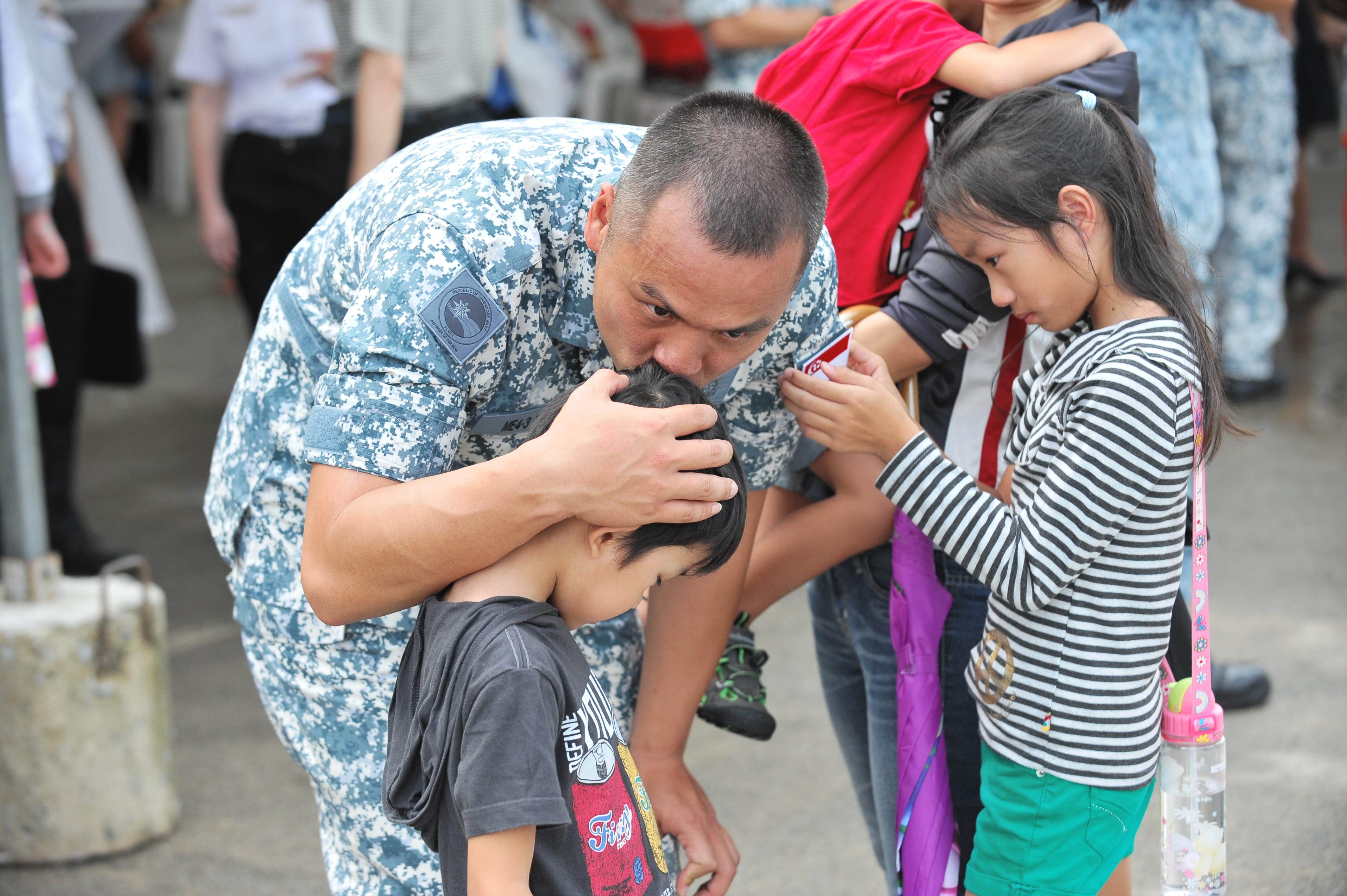A soldier hugs and kisses his child during a military homecoming event, with children in the background.