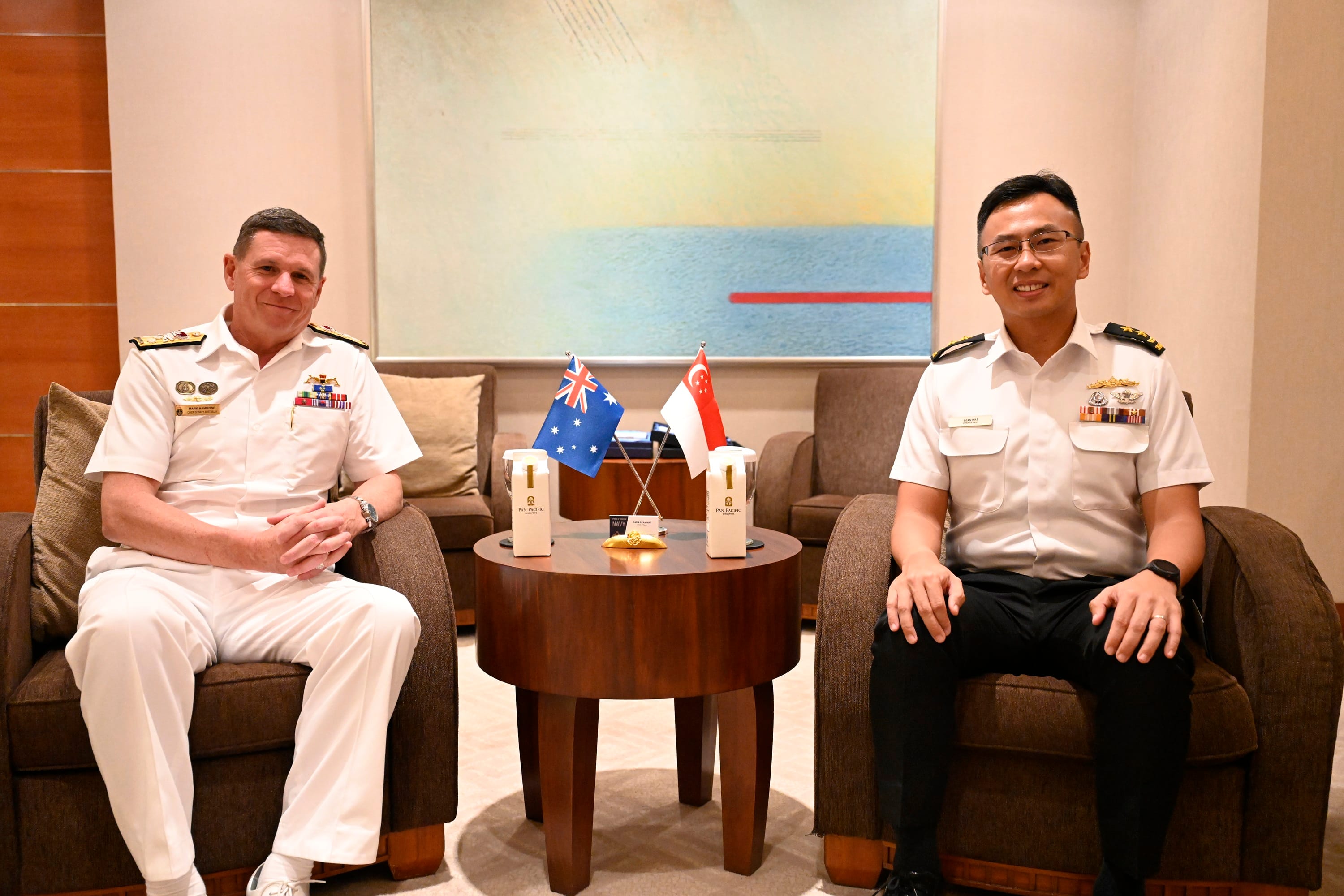 Two Navy officers in uniform sit in chairs with flags of Australia and Singapore on a table between them.