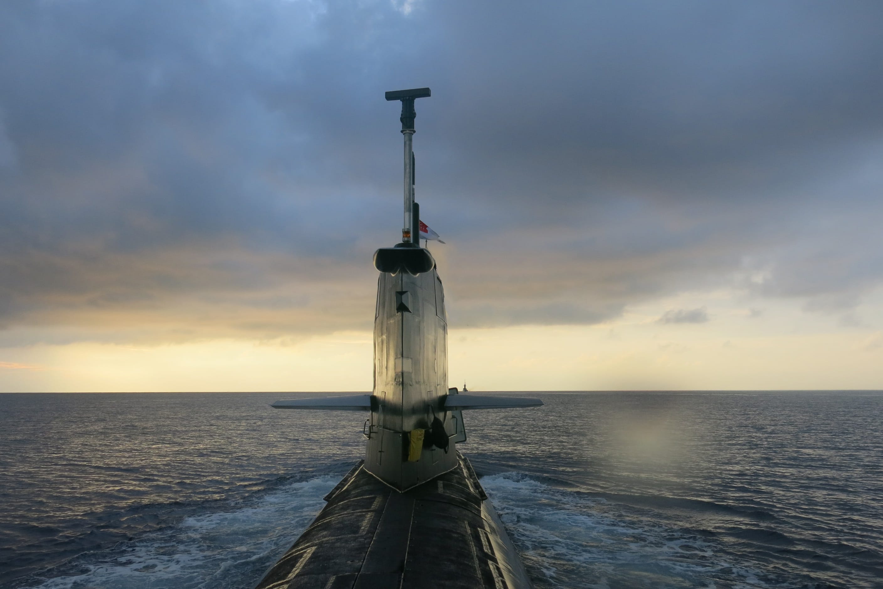 The rear view of a surfaced submarine with the Singapore flag on its mast under a cloudy sunset sky.