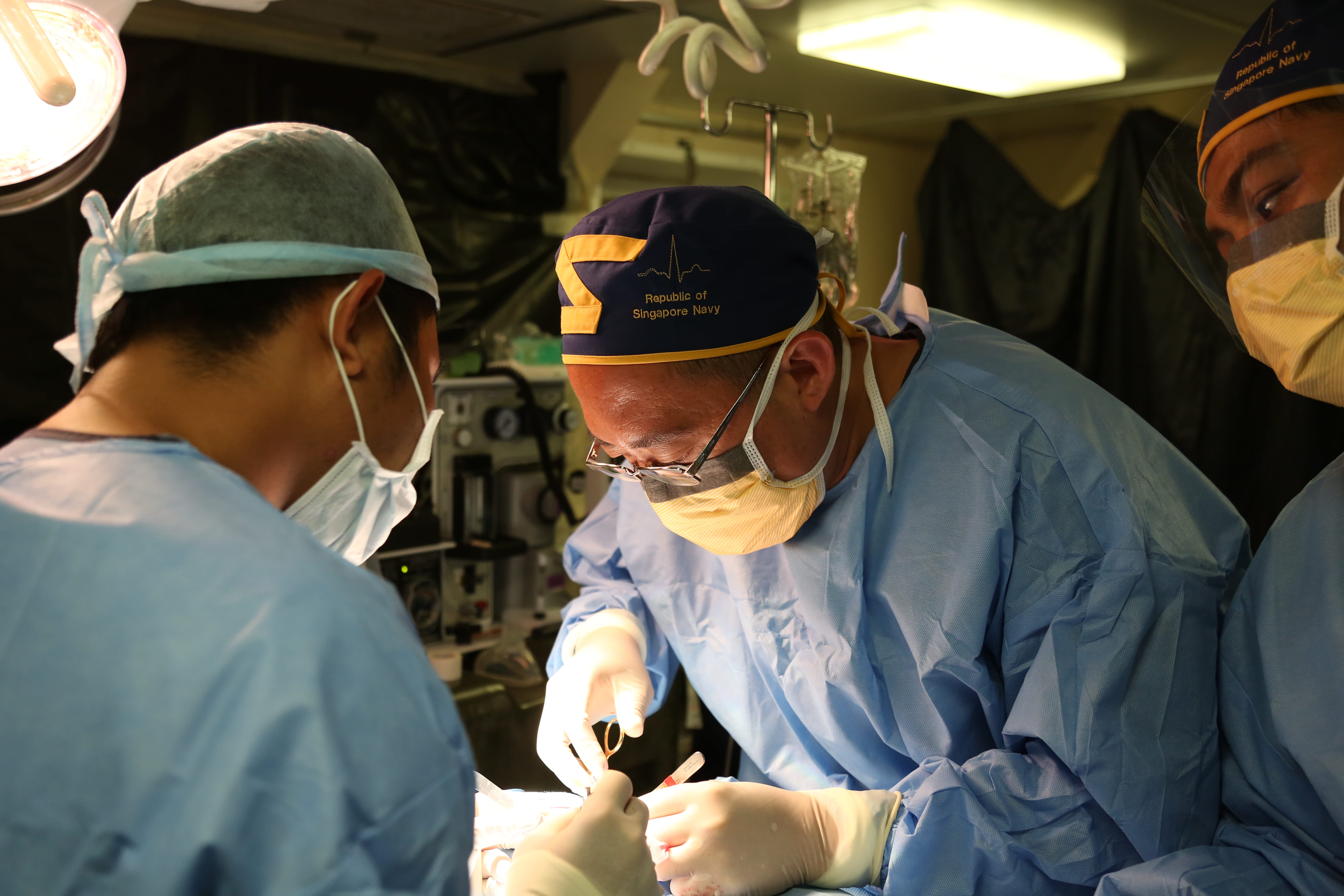 Naval surgeons perform a medical procedure in an onboard operating room, focused and wearing masks and gowns.